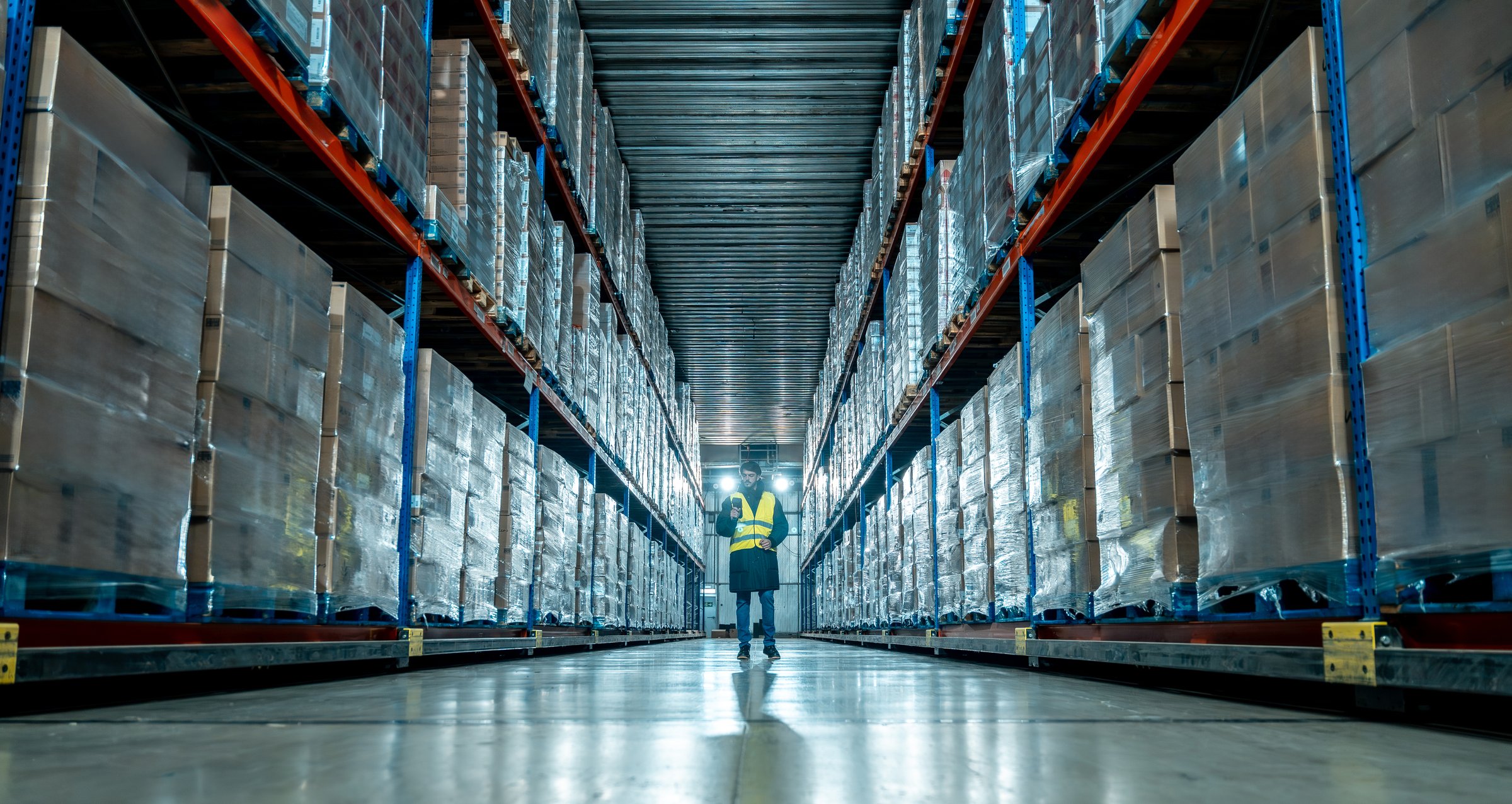 Worker wearing a high-visibility vest checking inventory and scanning packages in a vast cold storage warehouse aisle filled with stacked pallets of goods