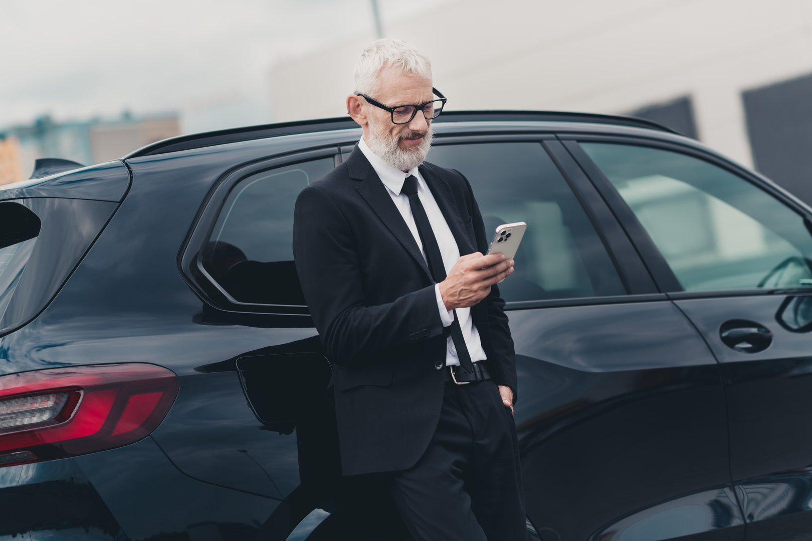 A mature businessman in formal attire stands beside a car in a city parking area, checking his smartphone. The image conveys travel, business, and modern technology concepts.