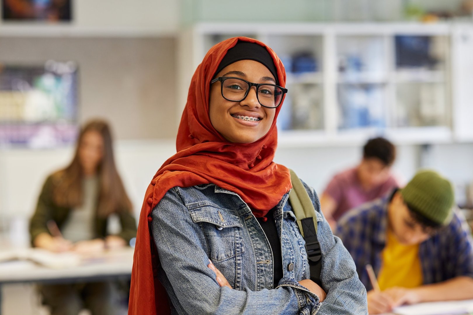 Portrait of happy and successful teen student smiles with a dental appliance in classroom with folded arms. Islamic girl wearing hijab and poses proudly in front of classmates during lesson. Portrait of proud and satisfied high school female student looking at camera.
