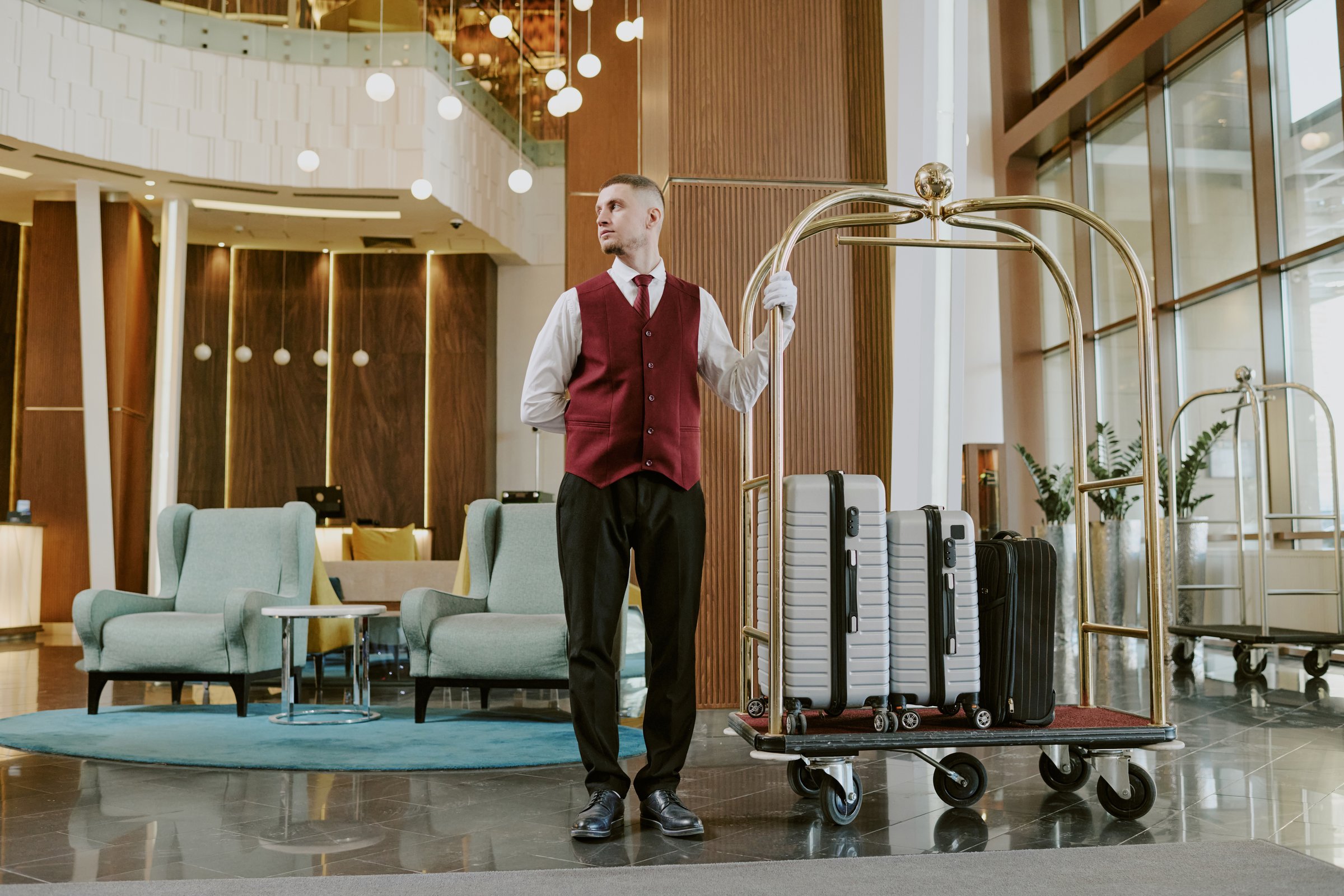 Low angle shot of young adult Caucasian bellboy standing near golden luggage cart in center of hotel lobby