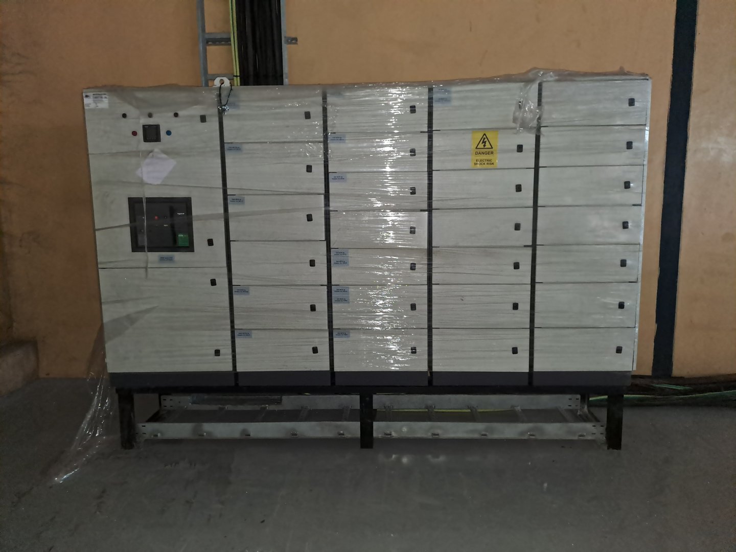 Electrical control panel with switches and knobs, wrapped in plastic, mounted on a metal frame against a beige wall.