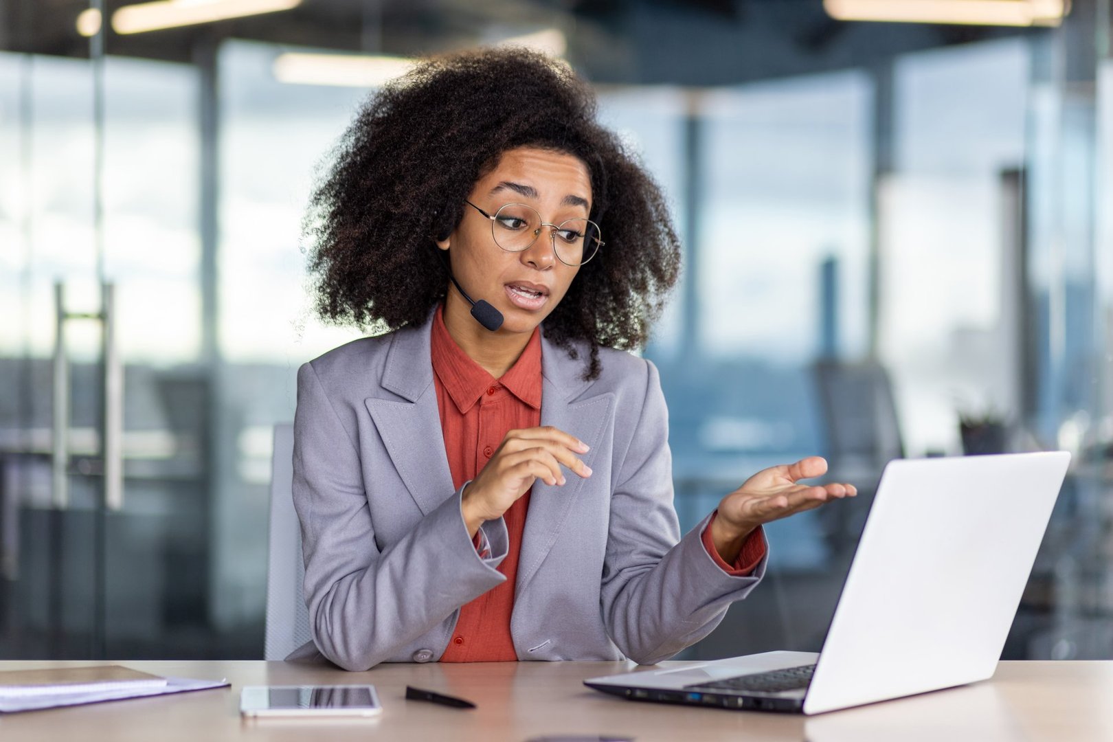 A young, professional woman with a curly afro wearing glasses and a headset is engaged in an animated video conference in a brightly lit, contemporary office setting, looking slightly puzzled or in need of clarification.