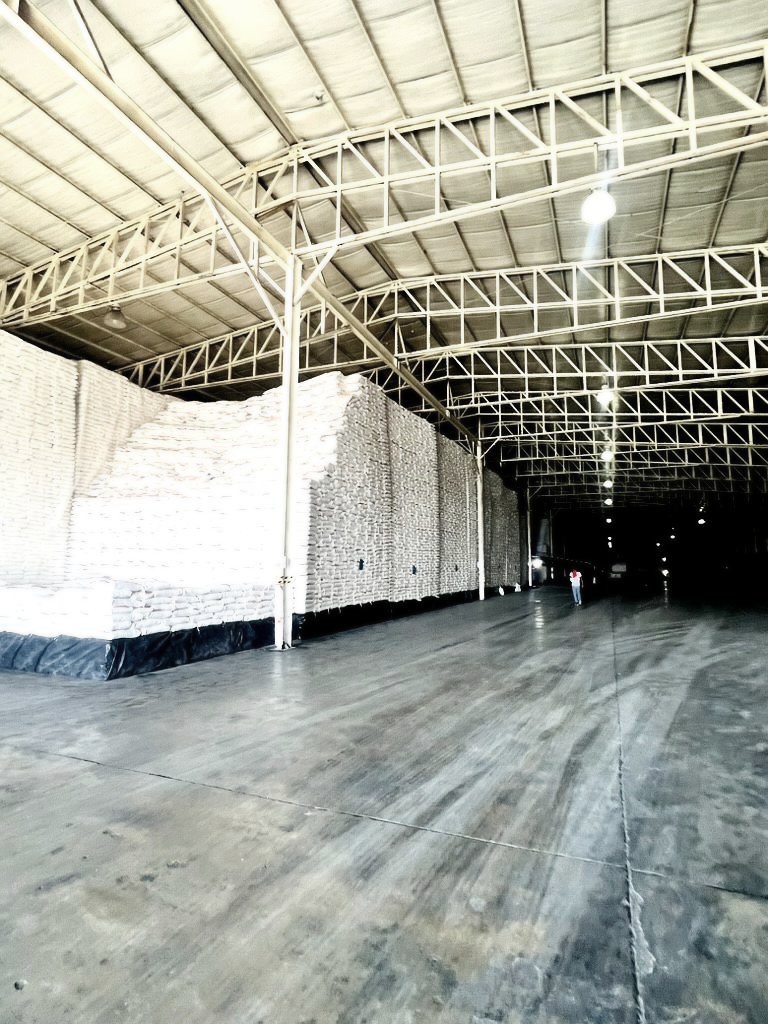 A large warehouse with stacks of packaged goods along the wall and a high ceiling with metal trusses.