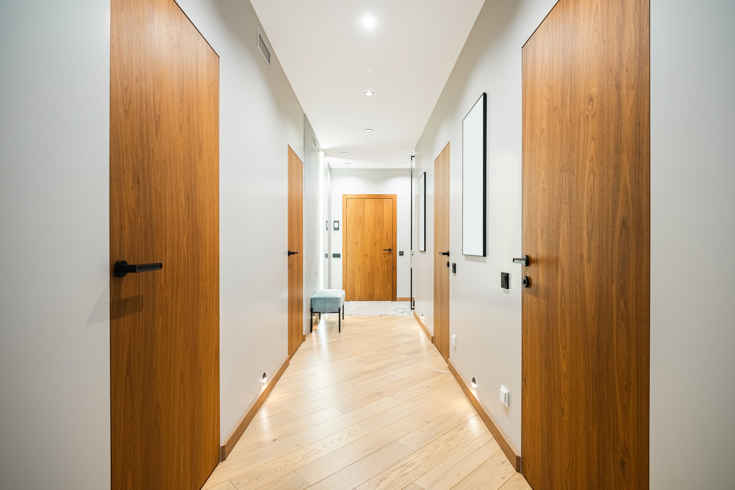 A spacious corridor showcases elegant wooden doors along its length, framed by smooth, neutral walls. Soft lighting highlights the clean lines and minimalistic design, creating a welcoming atmosphere.