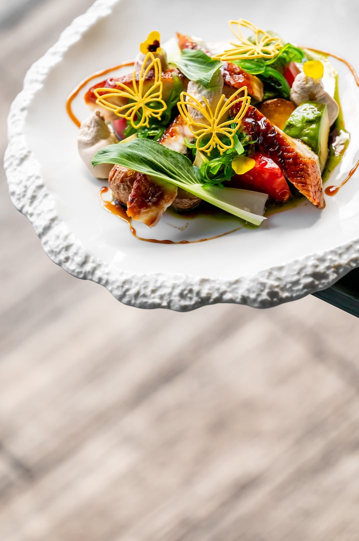 A gourmet dish with vibrant vegetables, fresh greens, succulent meat, and delicate yellow edible flowers, elegantly plated on a textured white plate with artistic sauce presentation