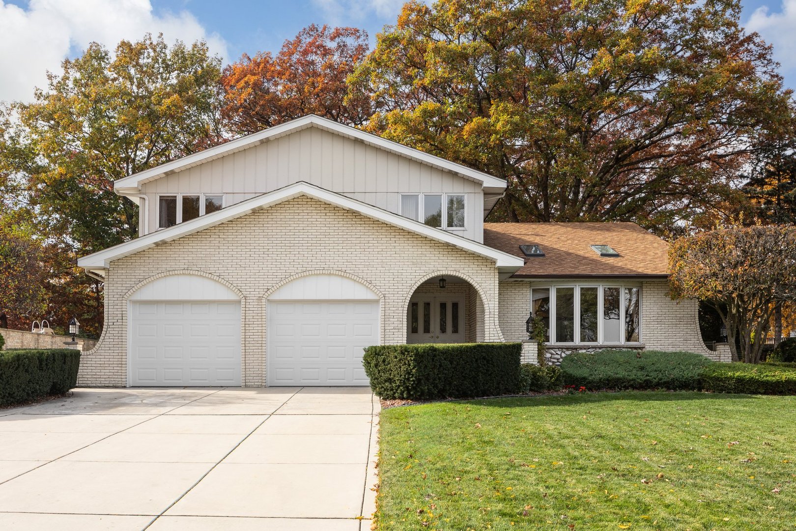 Geneva, IL, USA - October 30, 2024: A white brick two-story suburban house with a double garage, green lawn, and autumn trees