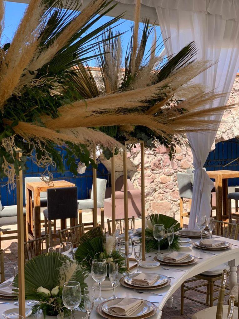 Elegant outdoor table setting with pampas grass decor, white plates, and folded napkins under a canopy.