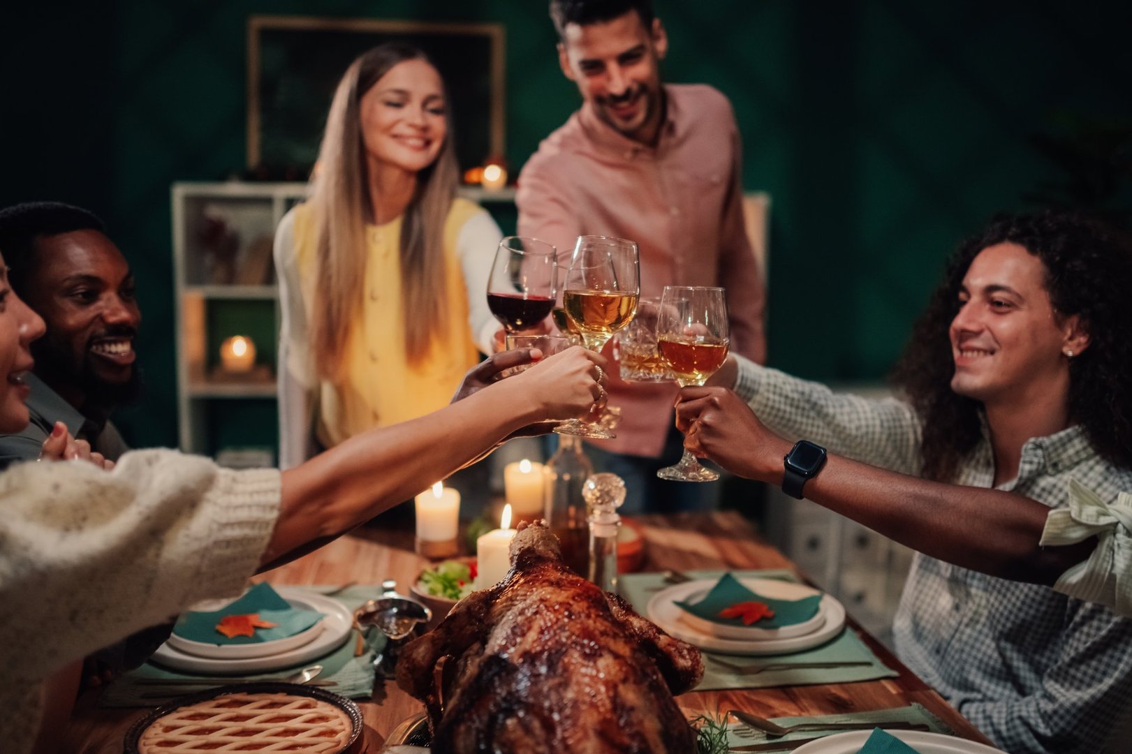 Group of friends raising wine glasses in a joyful toast while enjoying a festive thanksgiving dinner at home, surrounded by warmth and laughter