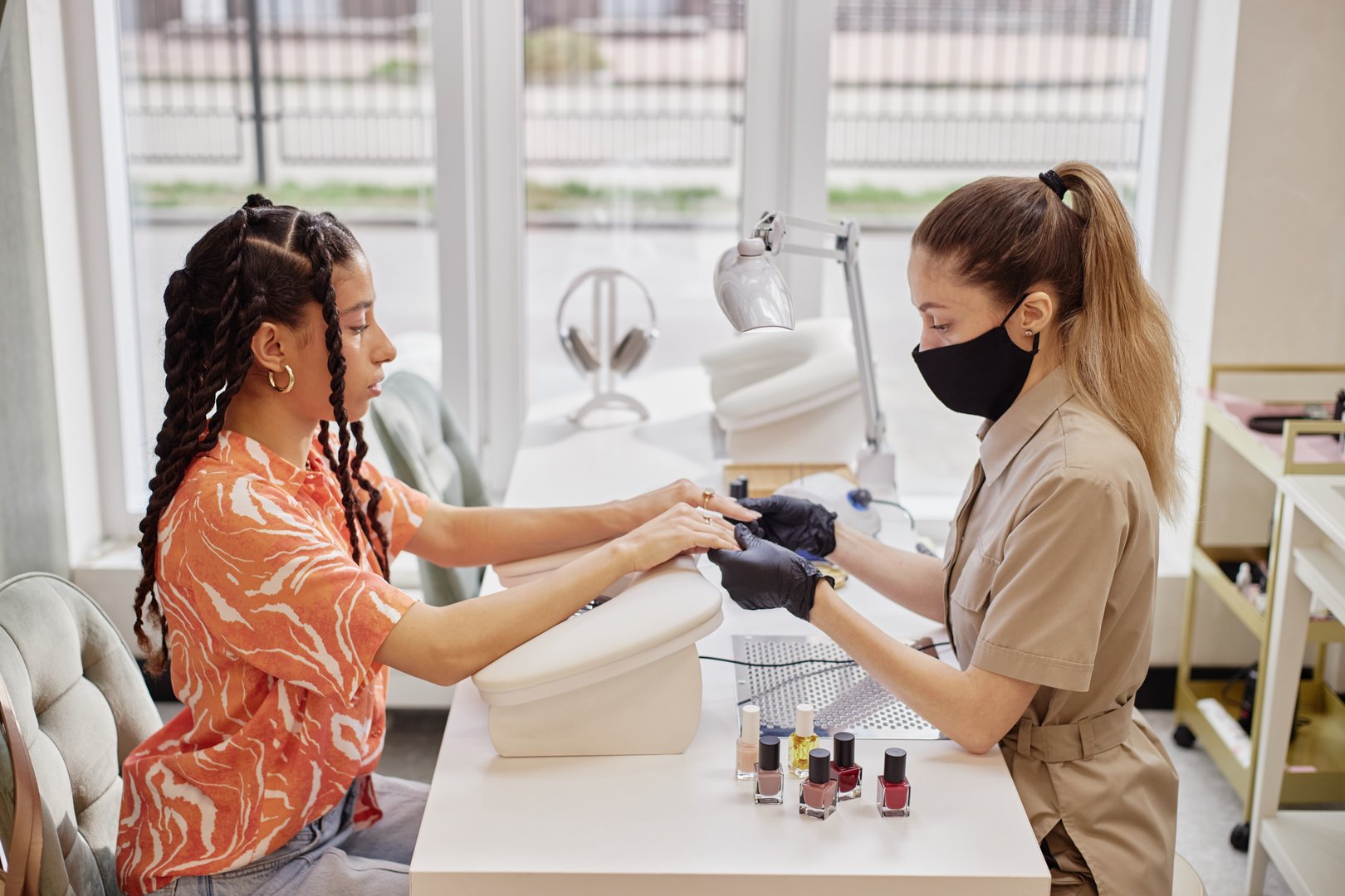 Biracial young adult woman receiving manicure from Caucasian middle aged woman wearing protective mask and gloves, both sitting at nail salon table with nail polish bottles visible