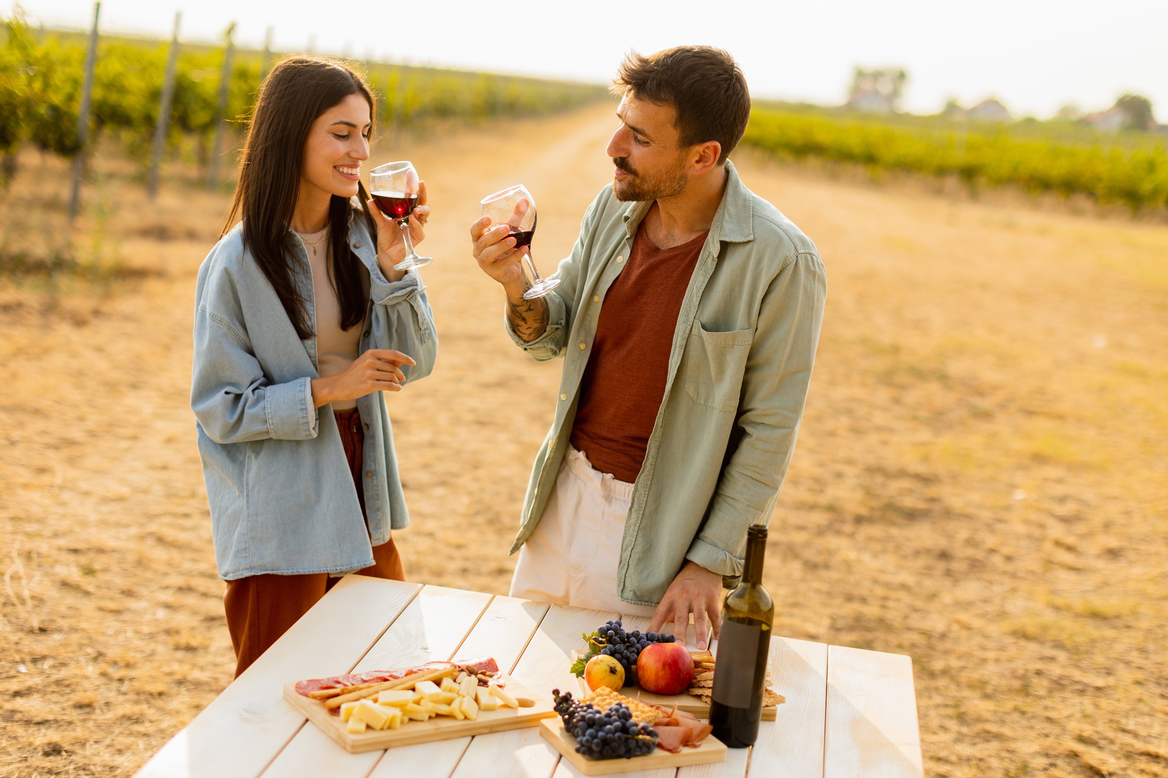 Couple savors red wine while standing by a rustic table filled with a variety of cheeses, fruits, and meats amidst a picturesque vineyard at dusk