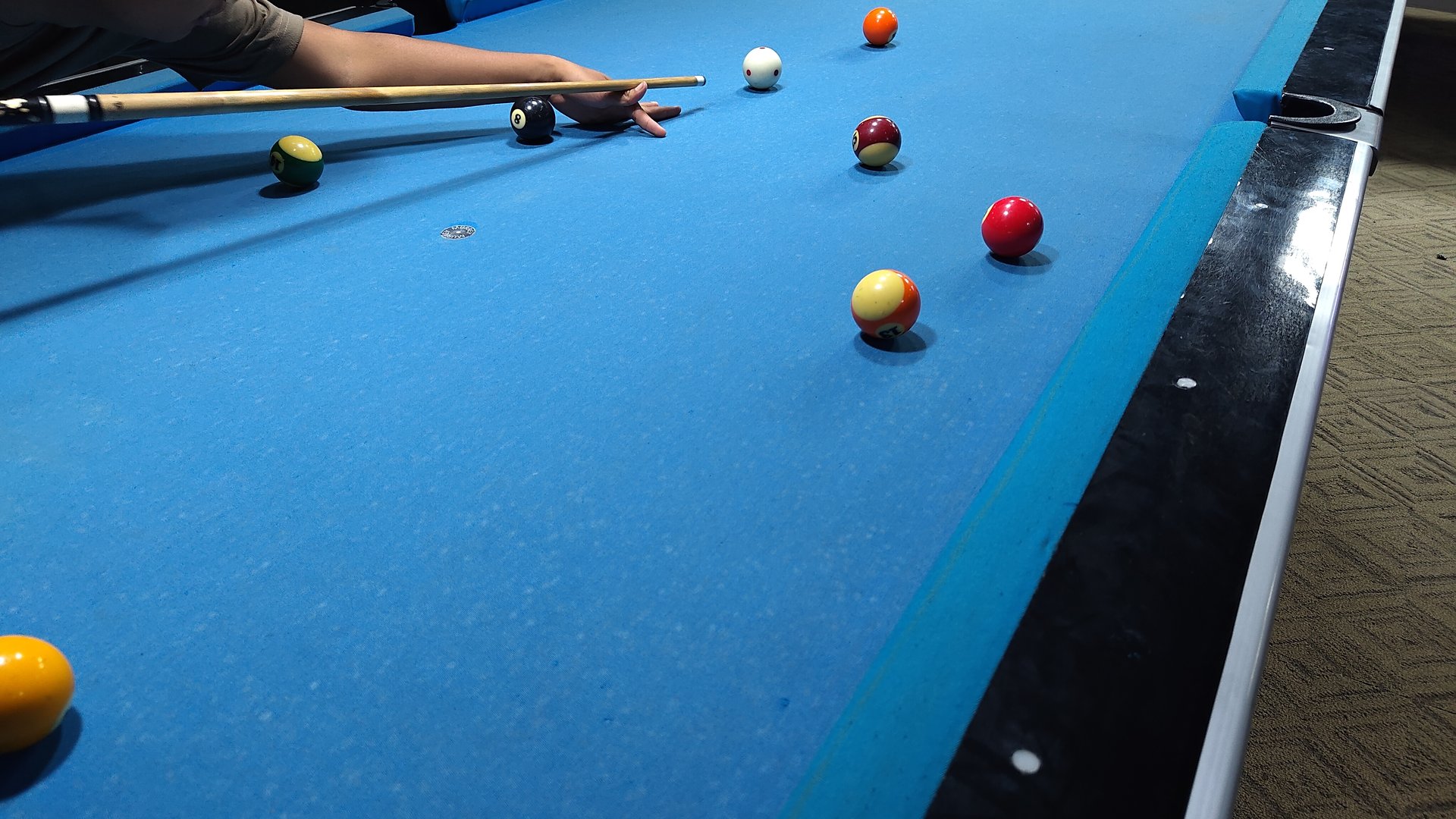 A billiards player lines up a shot on a blue pool table, with several colored balls spread across the felt. The scene captures focus, precision, and the casual leisure of indoor billiards.