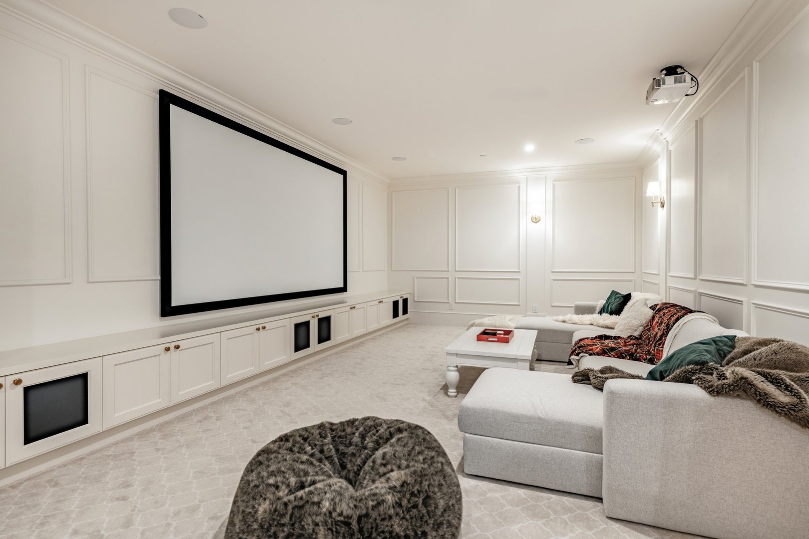 A home media theatre room with all white walls carpet sofa sectional and a very large projector and projection screen there are built in cabinets containing candy chips and an old fashioned style popcorn popper in red. There are colourful throw cushions blankets and pillows ont he sofa and a bean bag chair on the floor