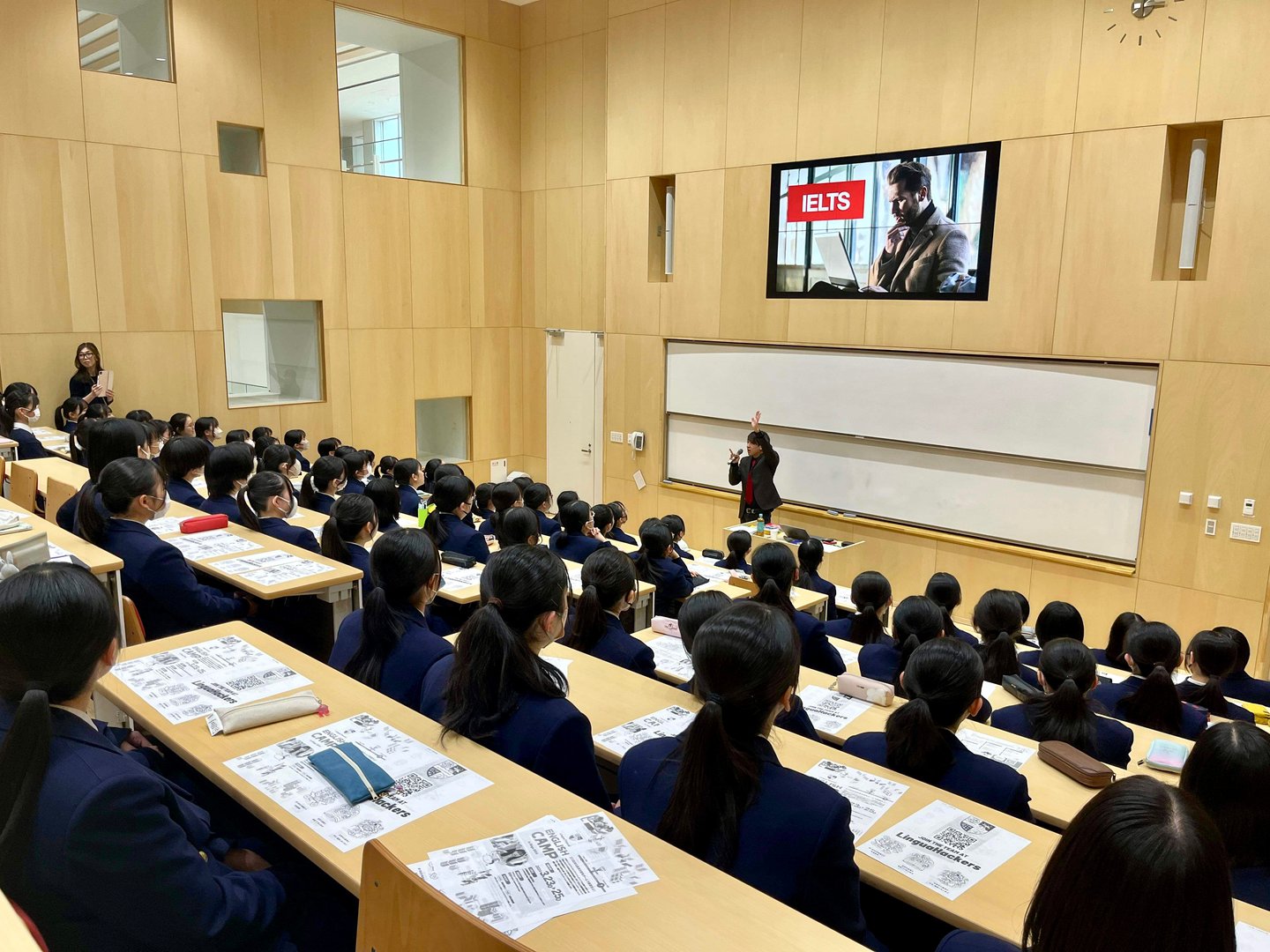 Students in uniforms seated in a lecture hall watching a presentation on IELTS displayed on a large screen.