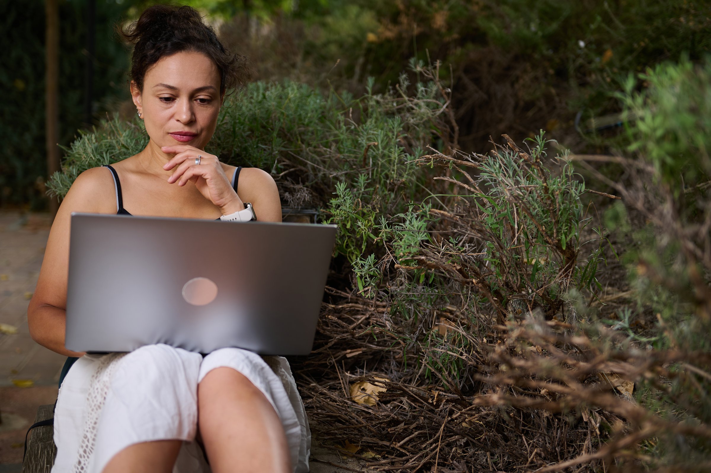 A woman sits outdoors amidst lush greenery, concentrating on her laptop. The warm lighting and serene surroundings complement her thoughtful expression, capturing a moment of digital focus.