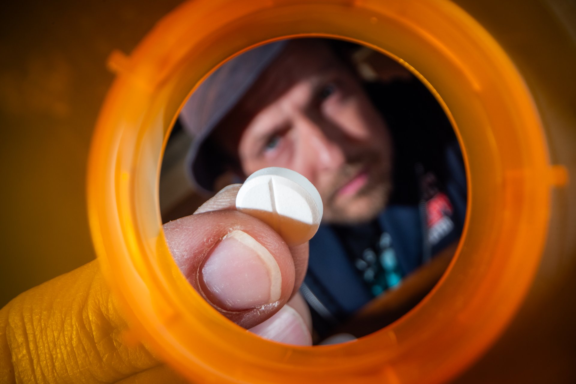 A powerful macro shot looking up from the inside of an empty, bright orange prescription pill bottle. The face of a troubled man is visible peering intensely down at the few remaining tablets.