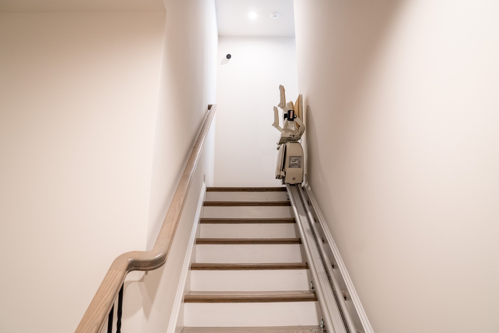 A stairlift installed on a narrow staircase with a wooden handrail, leading upward in a well-lit, white-walled space.