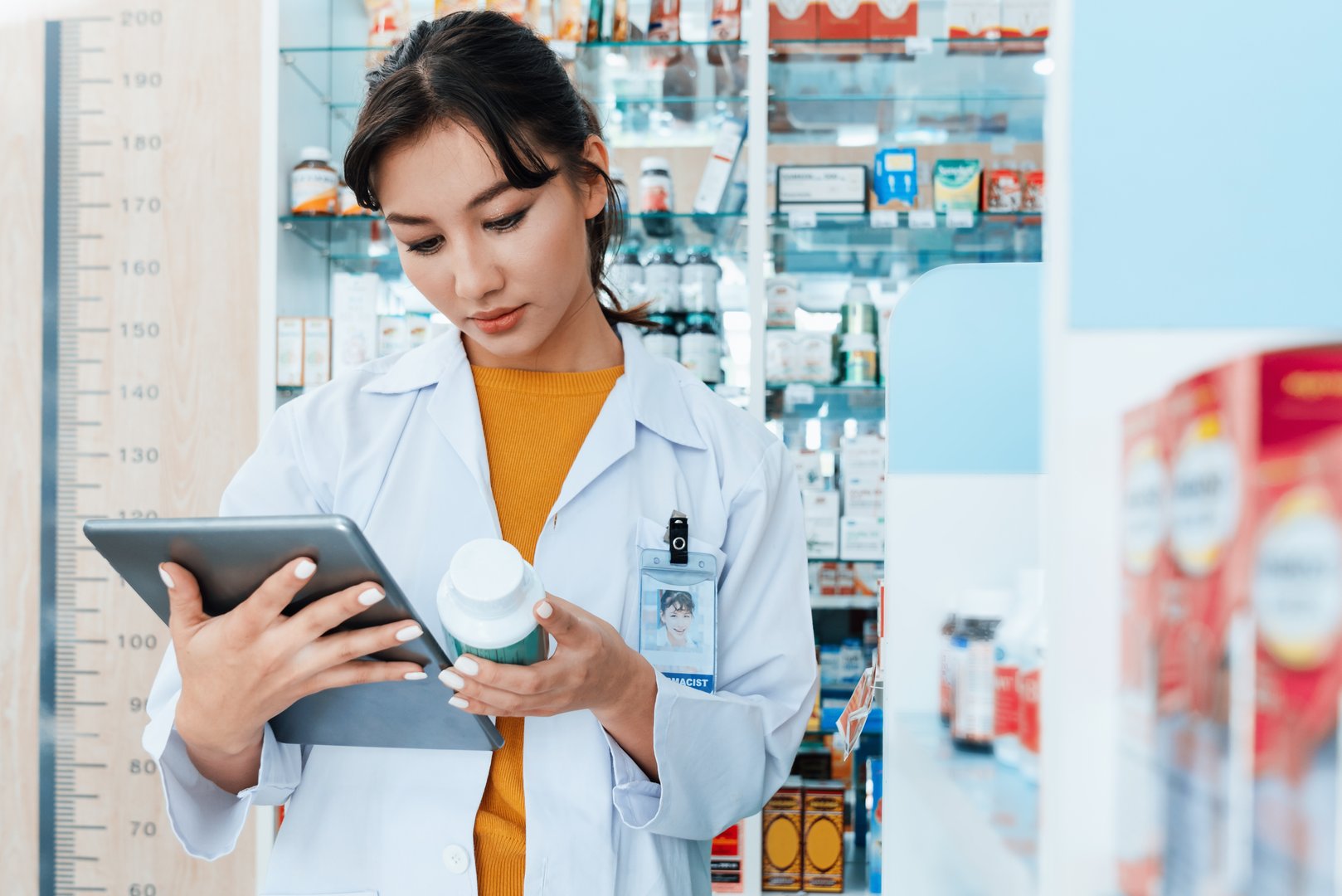 Portrait of a young asian female pharmacist and qualified pharmaceutical, medicine pill container or bottle mockup for copyspace at pharmacy. Drugstore concept with various medicine box in background.