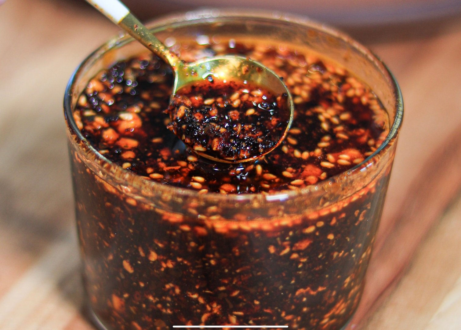 Close-up of a glass jar filled with spicy chili oil, with a spoon showing a mixture of chili flakes and sesame seeds.