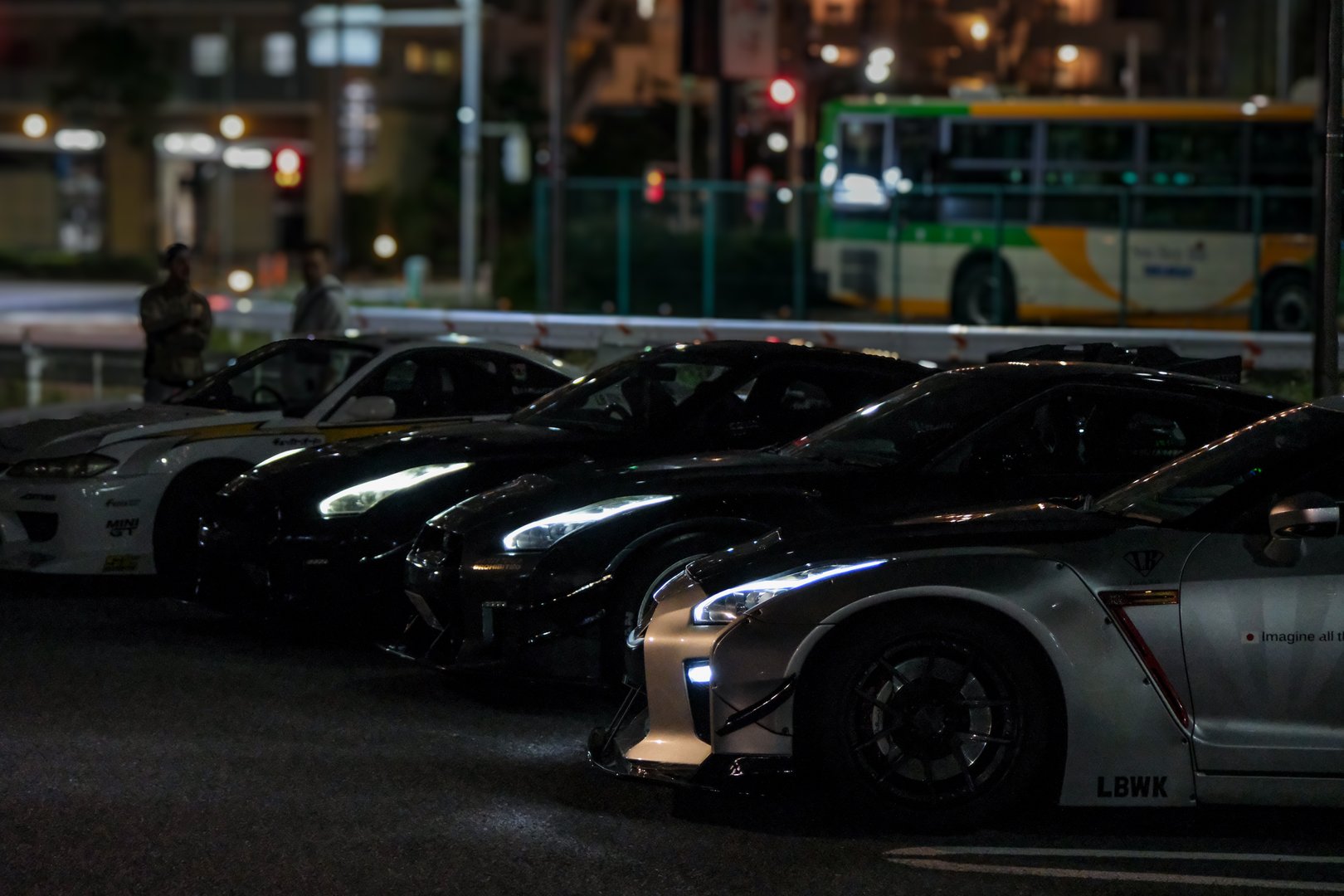 A lineup of high-performance sports cars is parked on a city street at night, illuminated by ambient lighting. The scene reflects urban car culture, showcasing modified vehicles and tuning style in a Japanese city