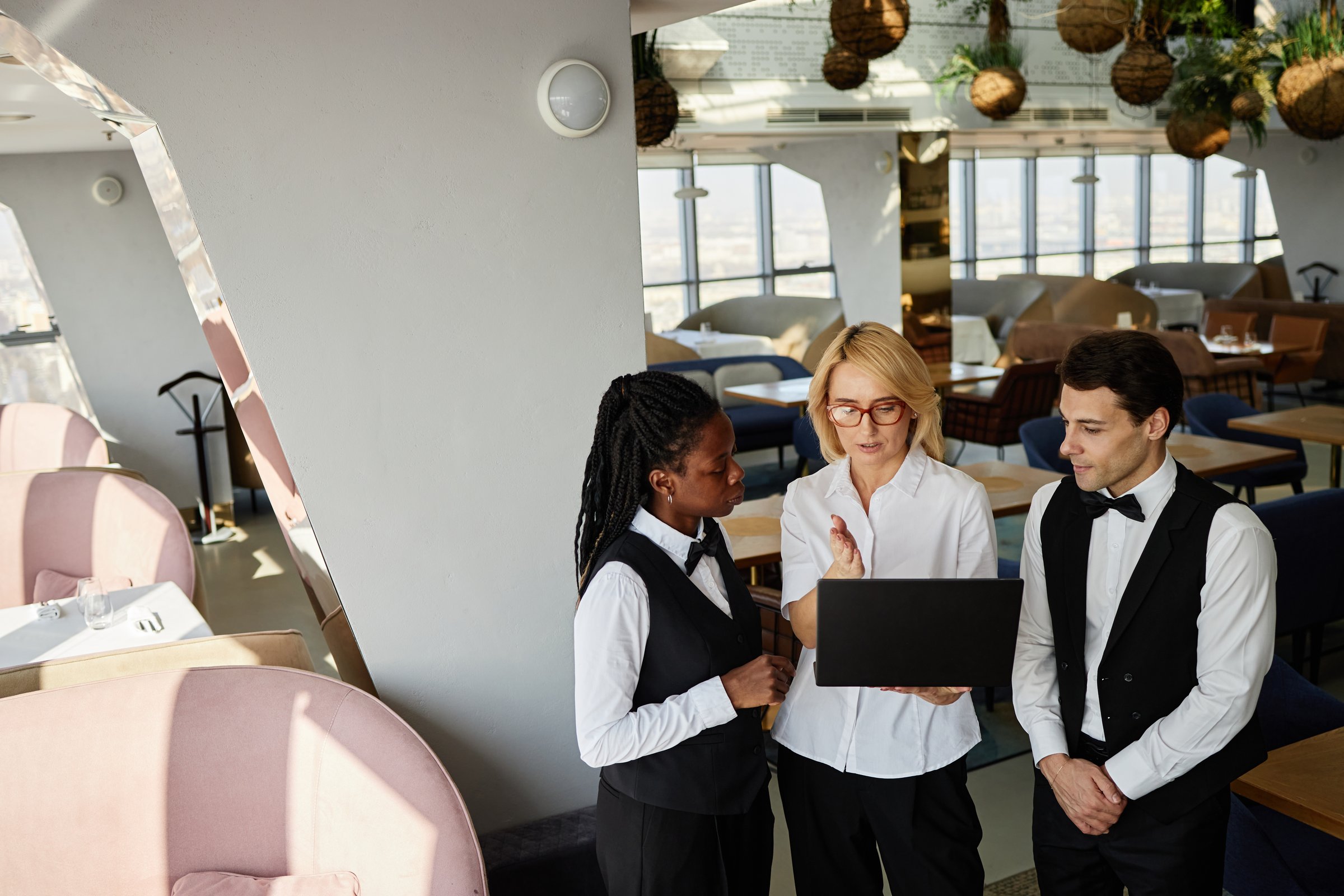 Middle aged Caucasian woman showing something on laptop to young adult Black woman and young adult Caucasian man in restaurant, all wearing uniforms and standing together