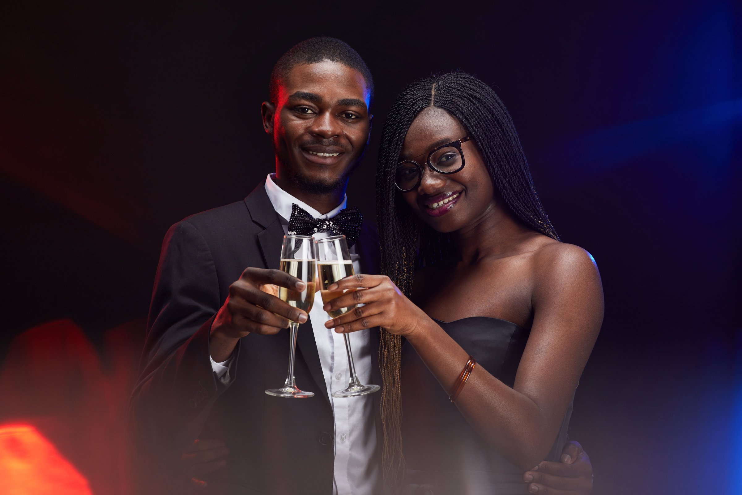 Waist up portrait of elegant African-American couple clinking champagne glasses while posing at party in dark