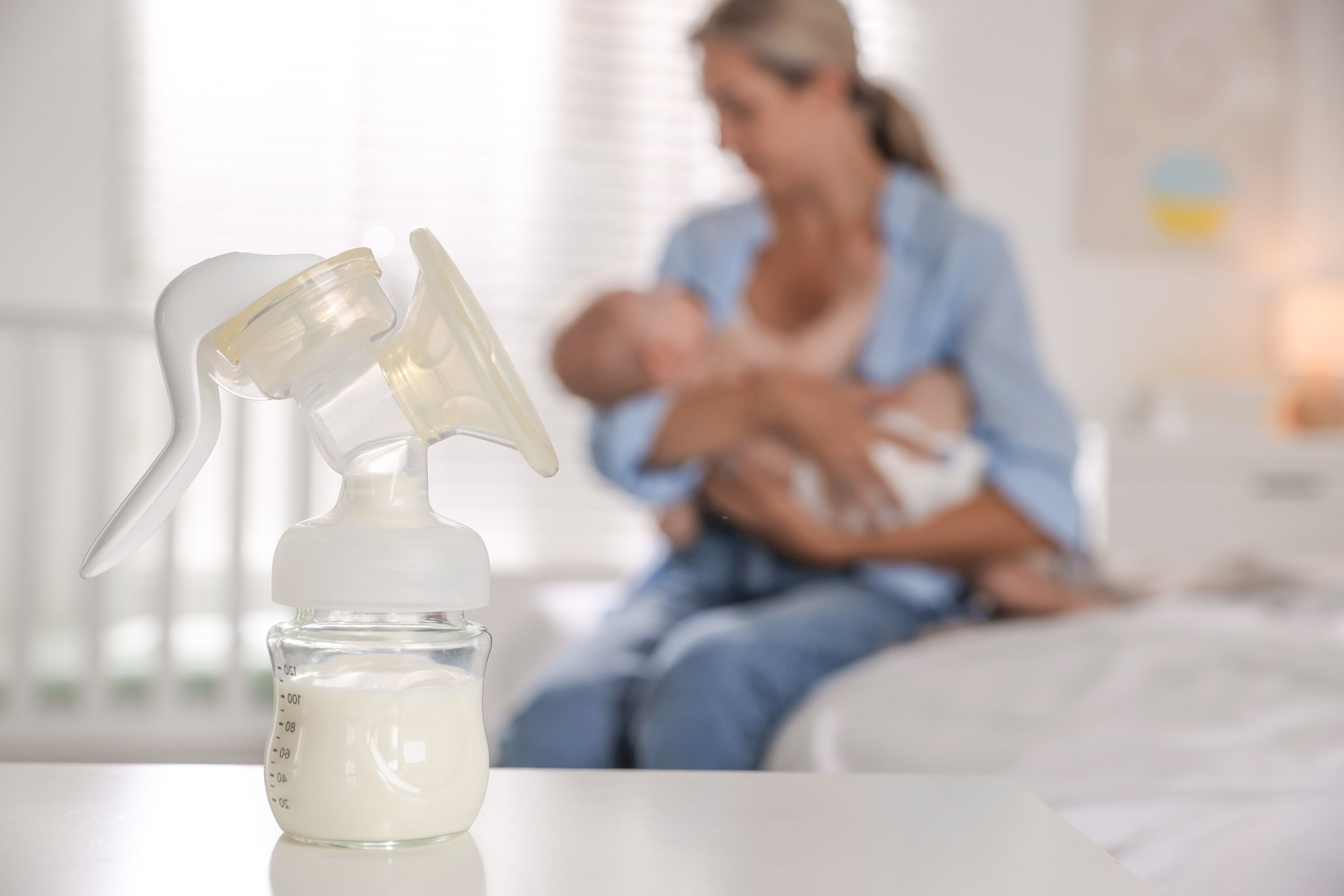 Mother holding her cute little baby indoors, focus on breast pump with milk