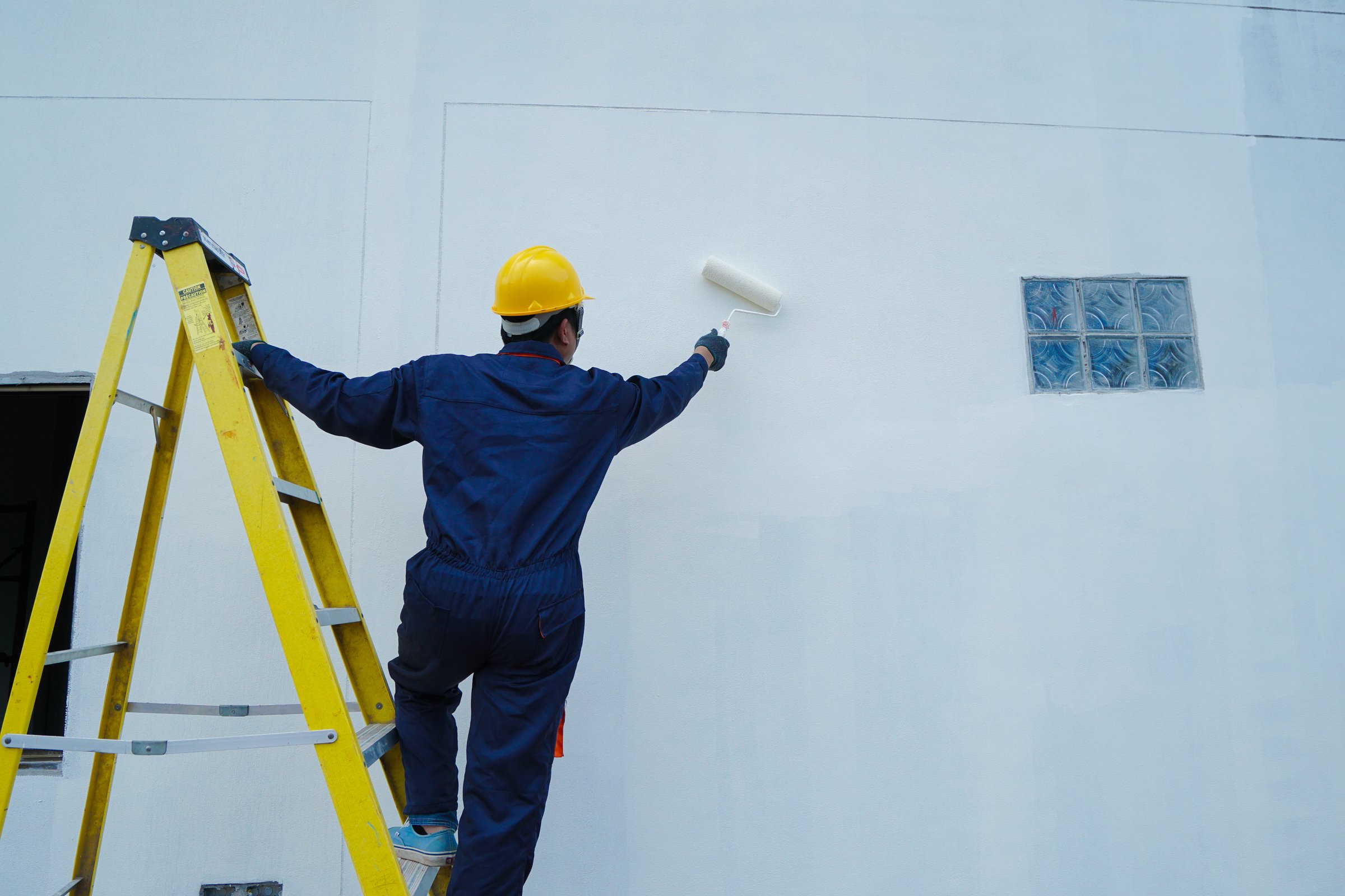 Construction workers climb a ladder while using a roller to paint a white house wall.