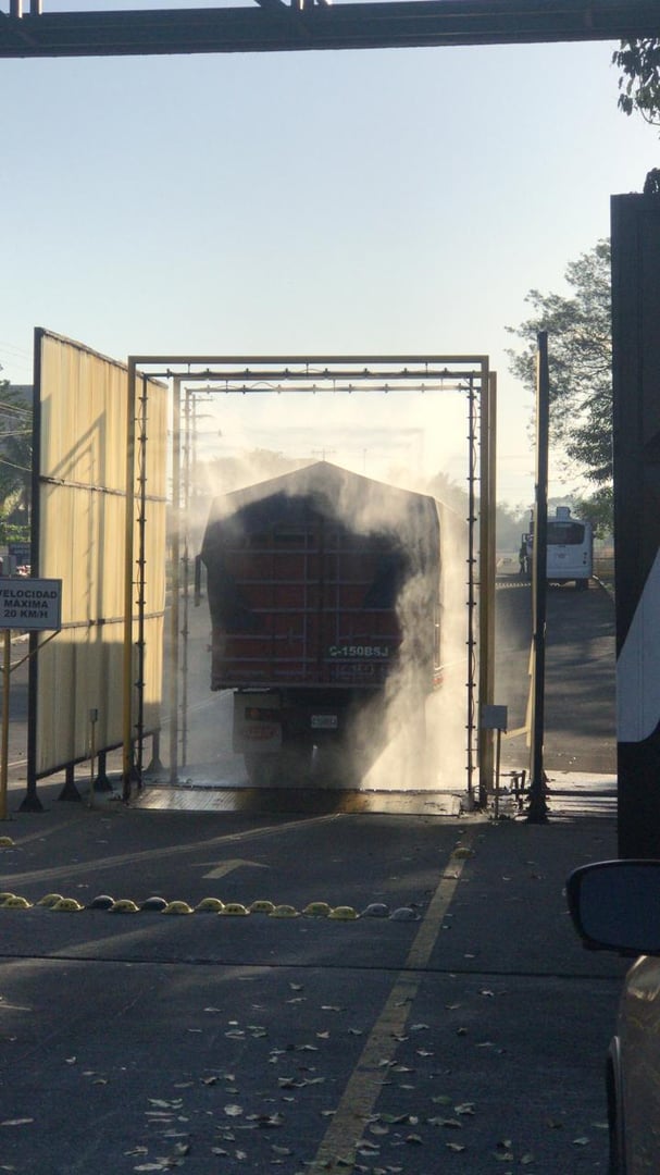 Truck passing through a vehicle sanitation booth, surrounded by mist, on a sunny day at a facility entrance.