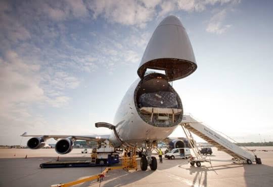 Cargo aircraft with open nose reveals interior loading area on tarmac, with stairs and ground crew nearby under a clear sky.