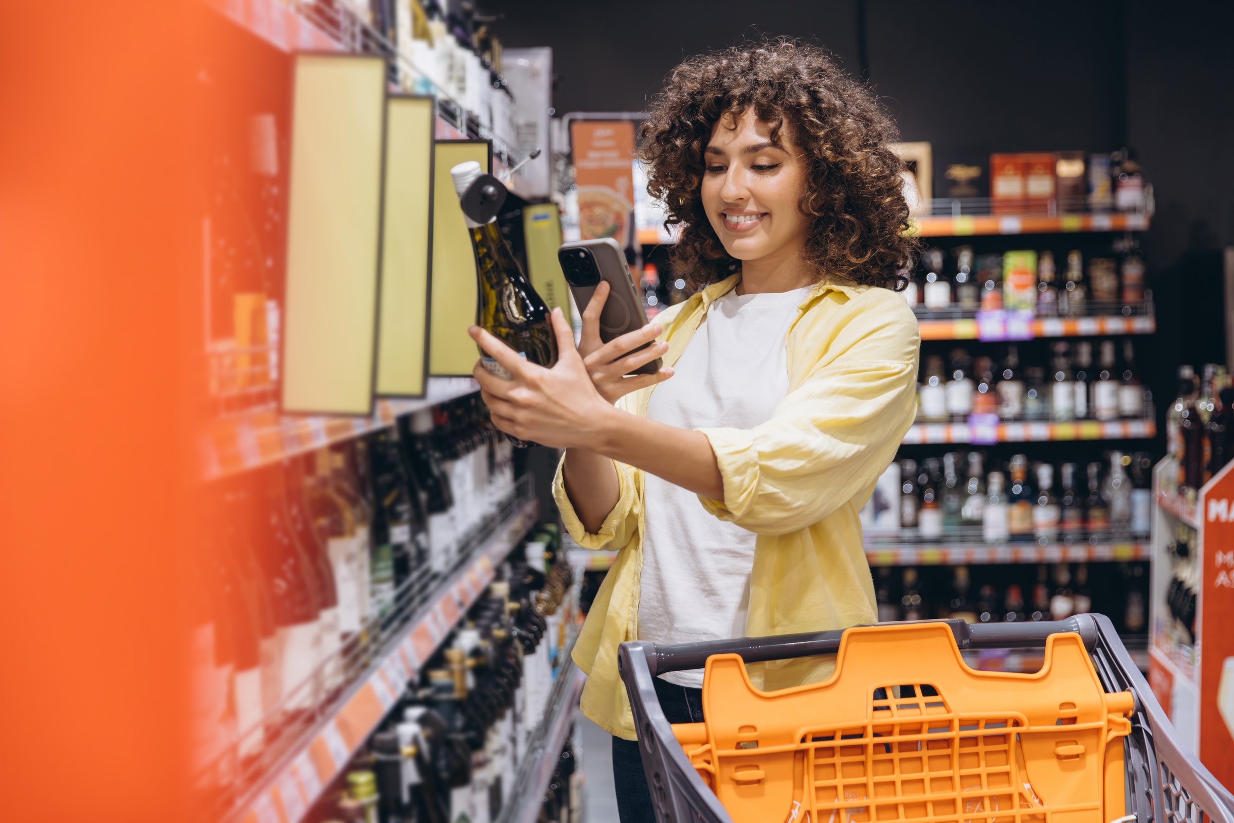 Smiling woman using phone to scan product information of wine bottle while shopping in supermarket