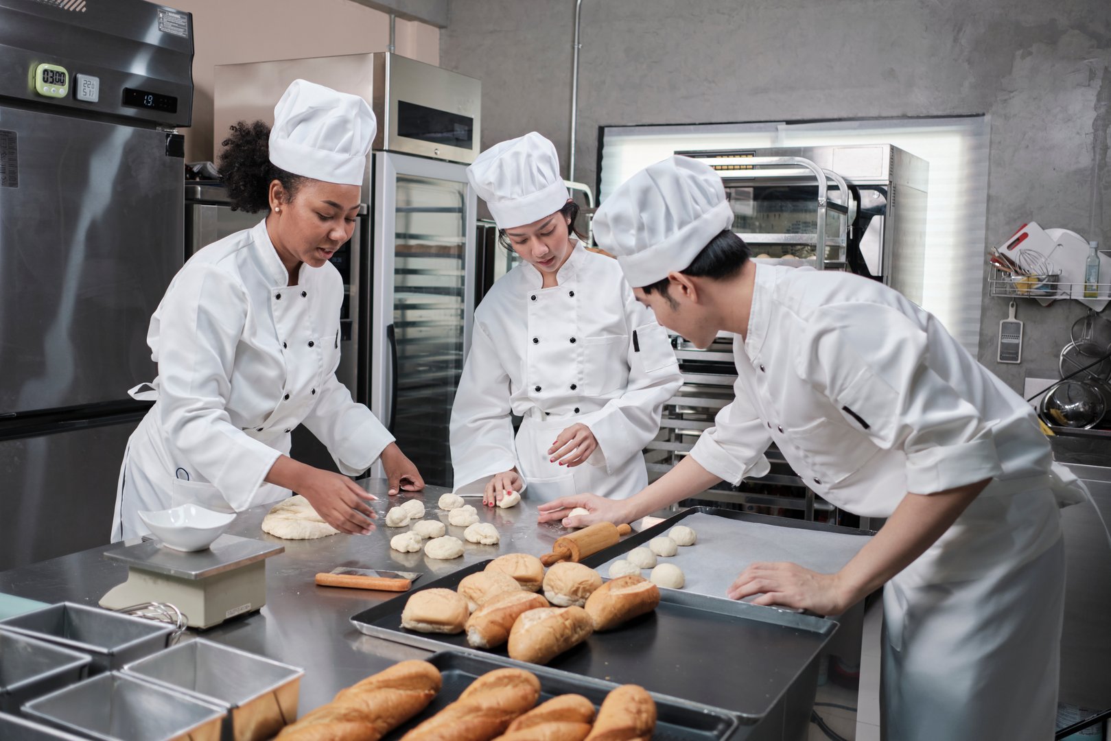 Professional gourmet team, three young chefs in white cook uniforms and aprons knead pastry dough and eggs, prepare bread, and fresh bakery food, baking in oven at stainless steel restaurant kitchen.