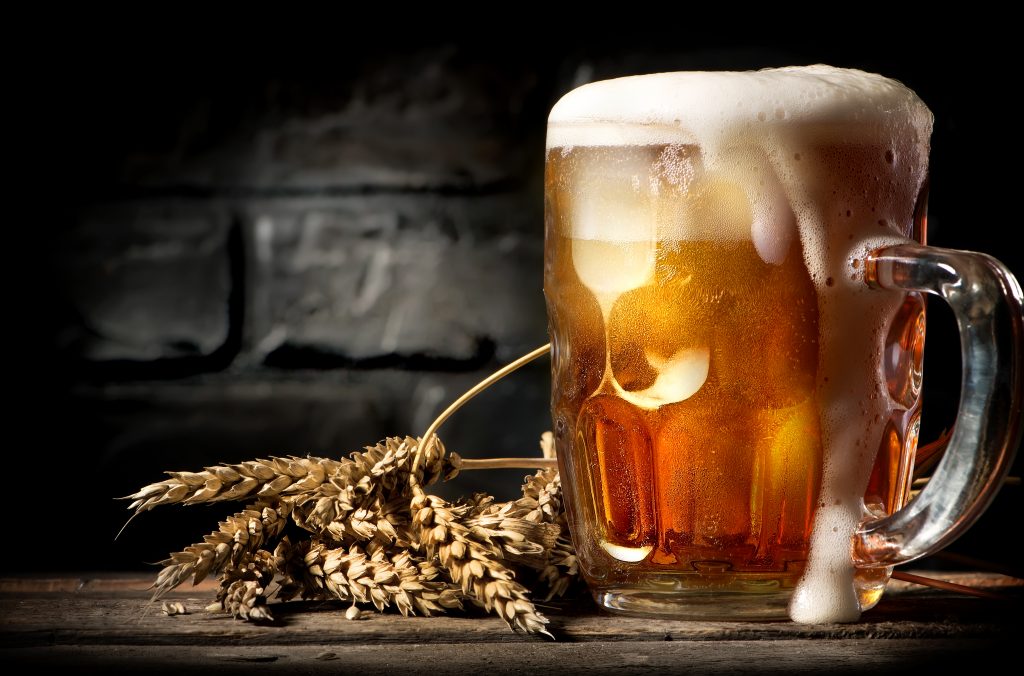 A frothy mug of beer with wheat stalks on a wooden table against a dark brick background.