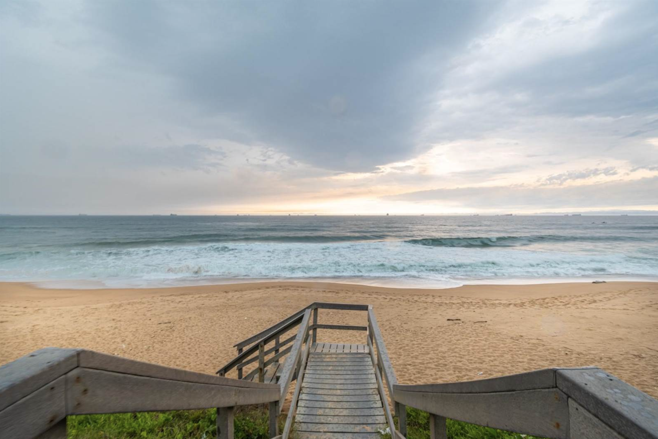 Wooden stairs leading to a sandy beach with ocean waves and a cloudy sky at sunrise or sunset.