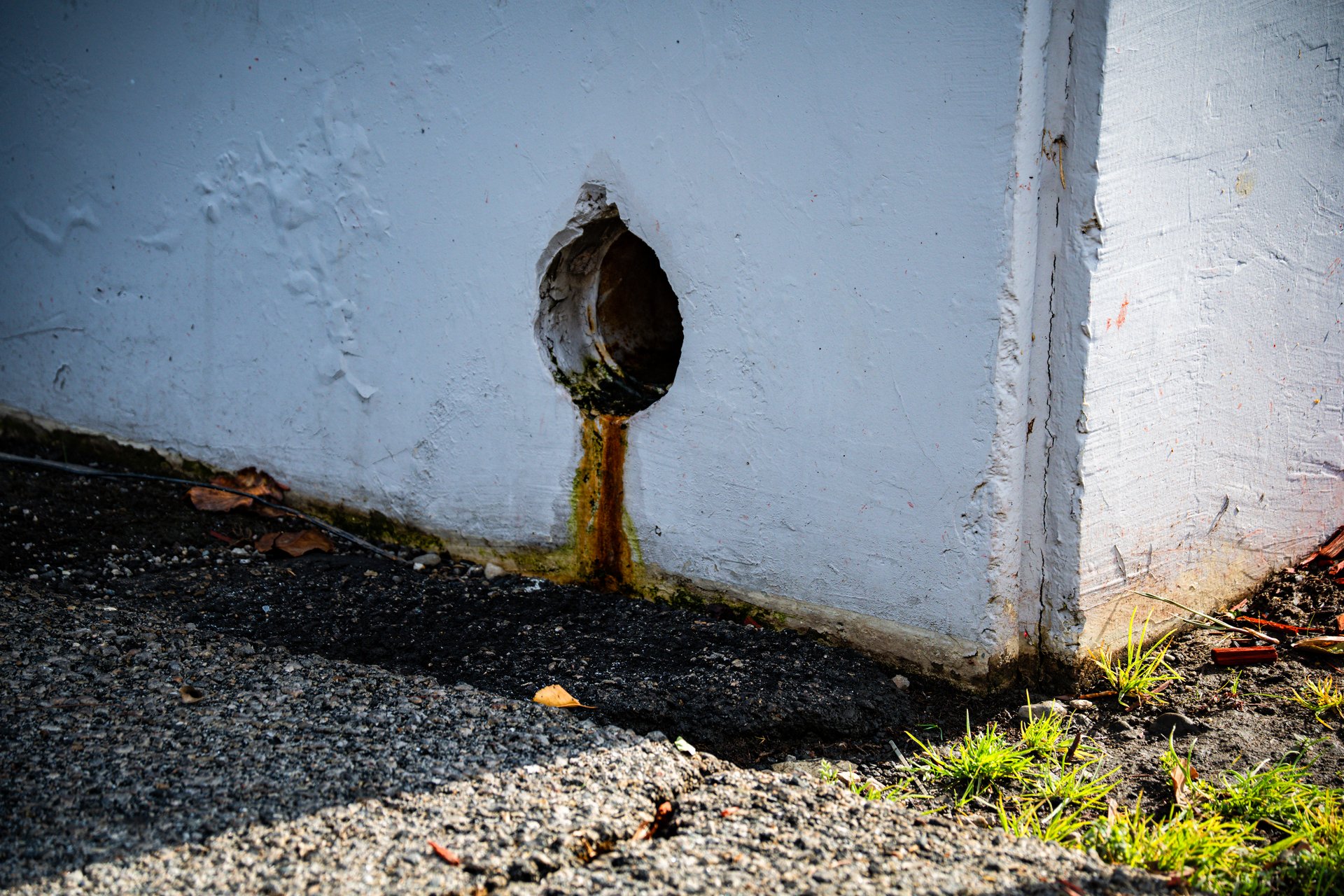 A close-up view of a weathered drainpipe on a building exterior, showing signs of rust and peeling paint, with gravel and grass at its base