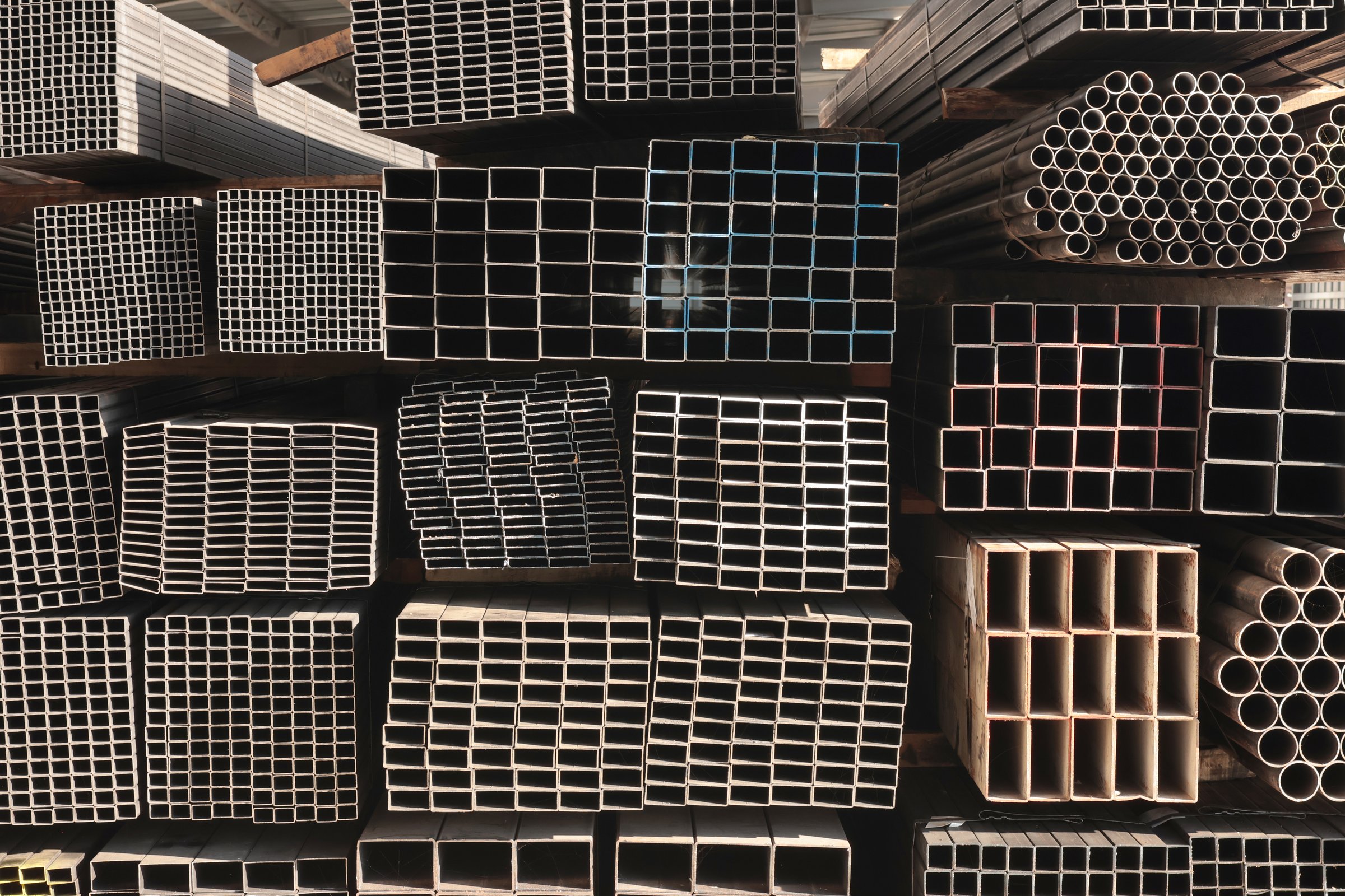 Close-up of stacked square steel hollow section profiles and round steel pipes with visible cut ends stored inside an industrial metal warehouse.