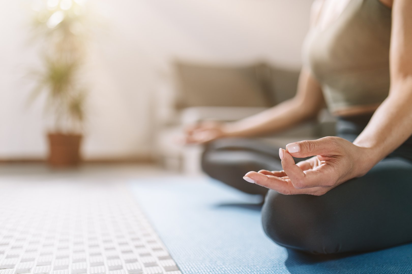 A person sits cross-legged on a yoga mat, hands positioned in a meditation mudra. The bright room features soft lighting and indoor plants, creating a calming atmosphere for mindfulness.