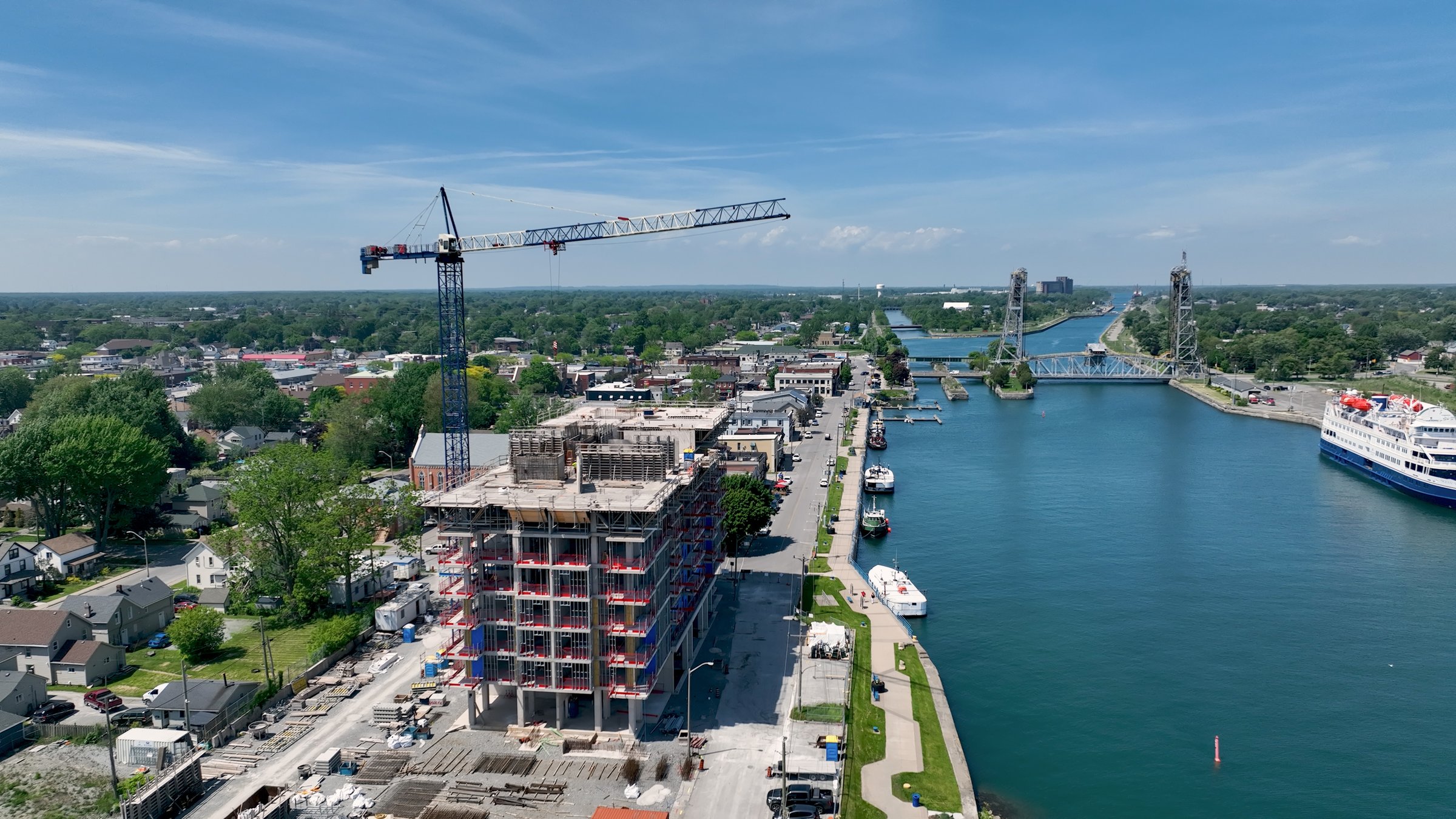 Captured on a clear, sunny day, a blue construction crane works on a new condo building under construction in an urban city located next to a canal.