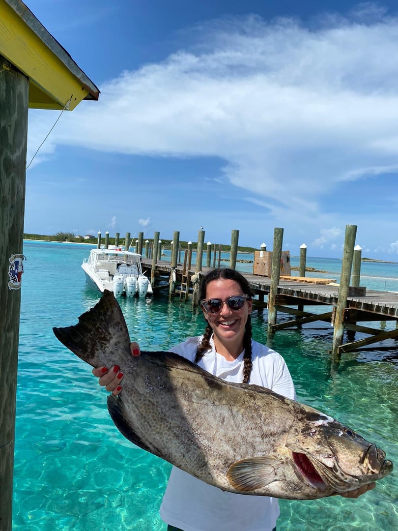 Person holding a large fish near a dock with clear turquoise water and boats in the background.