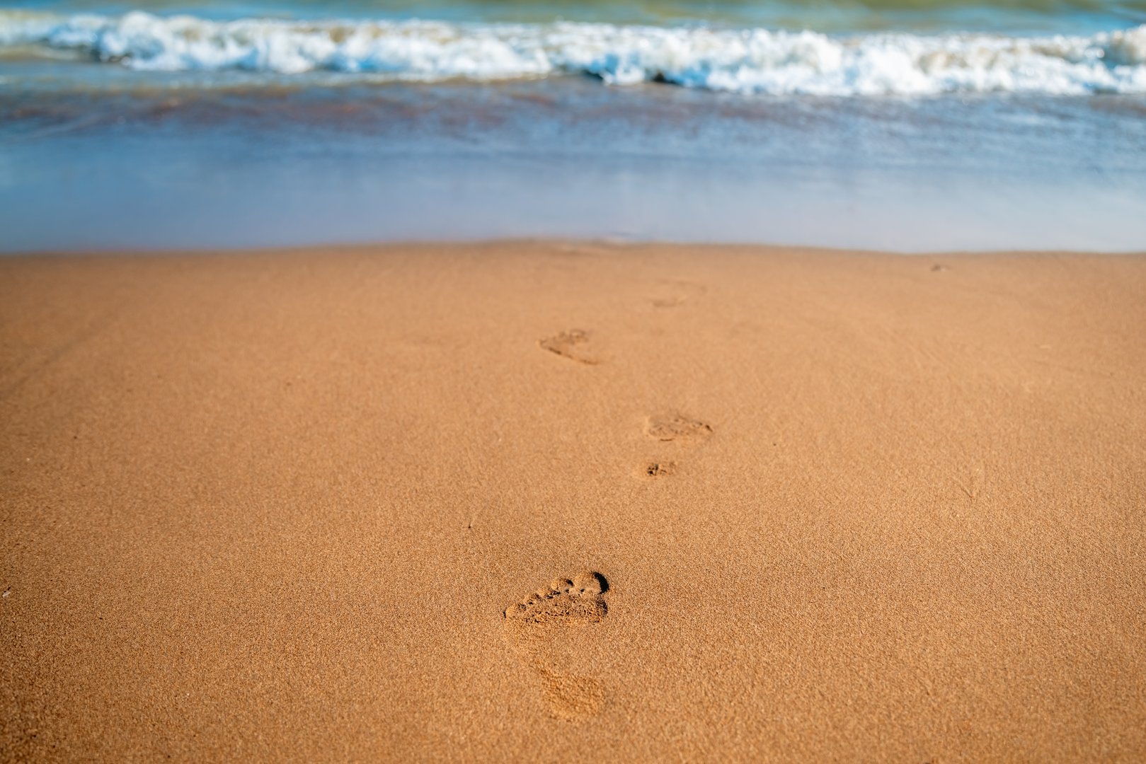 Footprints in the sand stretch away from the viewer, leading toward gentle ocean waves on a sunlit beach, creating a peaceful atmosphere of solitude and serene tranquility