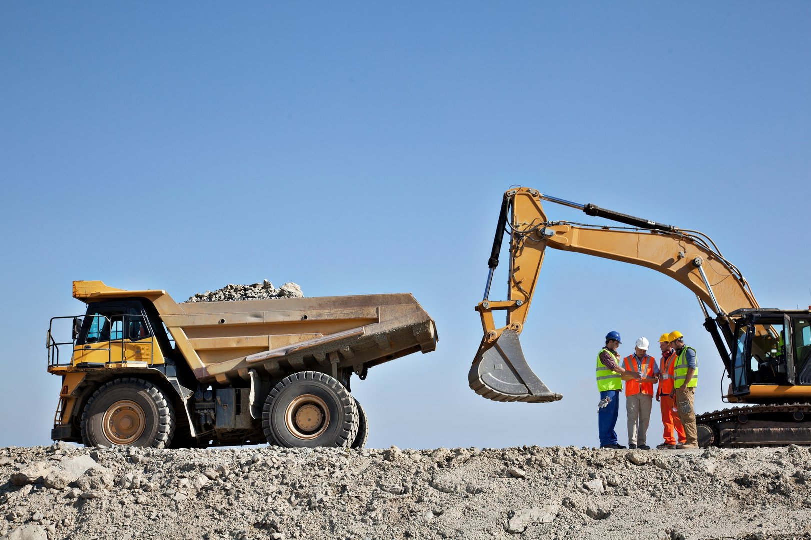 Construction workers in helmets and vests discussing near an excavator and a dump truck on a dirt site under a clear sky.