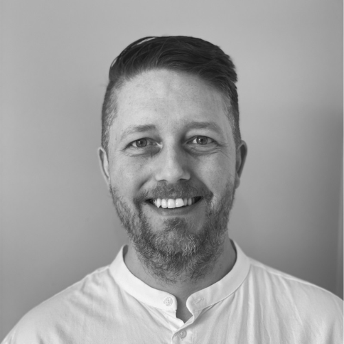 Smiling man with a beard and a short hairstyle wearing a white shirt against a plain background.