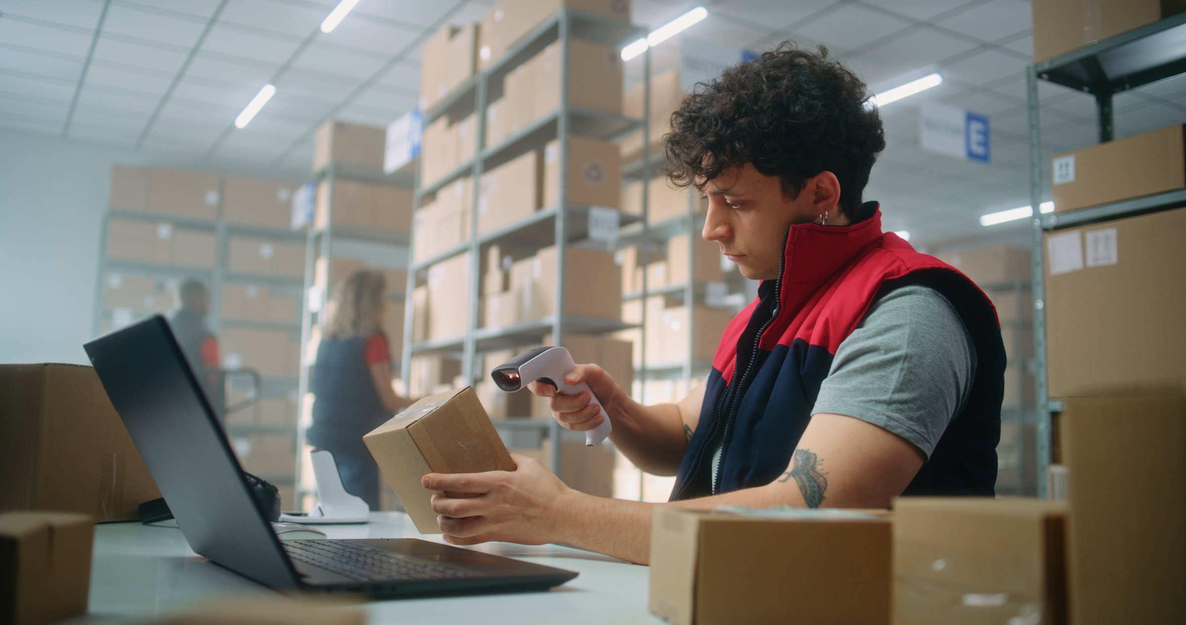 Parcel processing specialist scans package using barcode scanner, prepares for shipping to customer, works on laptop. Warehouse employees carrying boxes. E-commerce store or postal service storage.