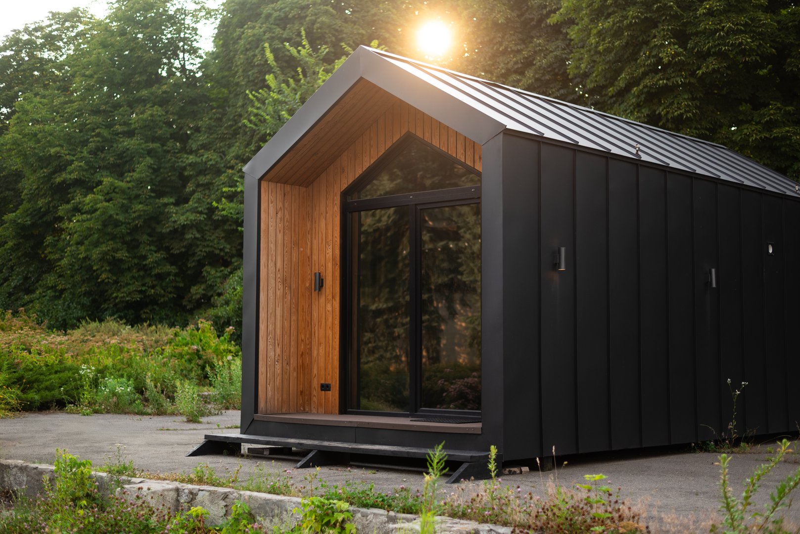 Modern tiny house with wooden and black metal facade, surrounded by greenery, lit by soft evening sunlight.