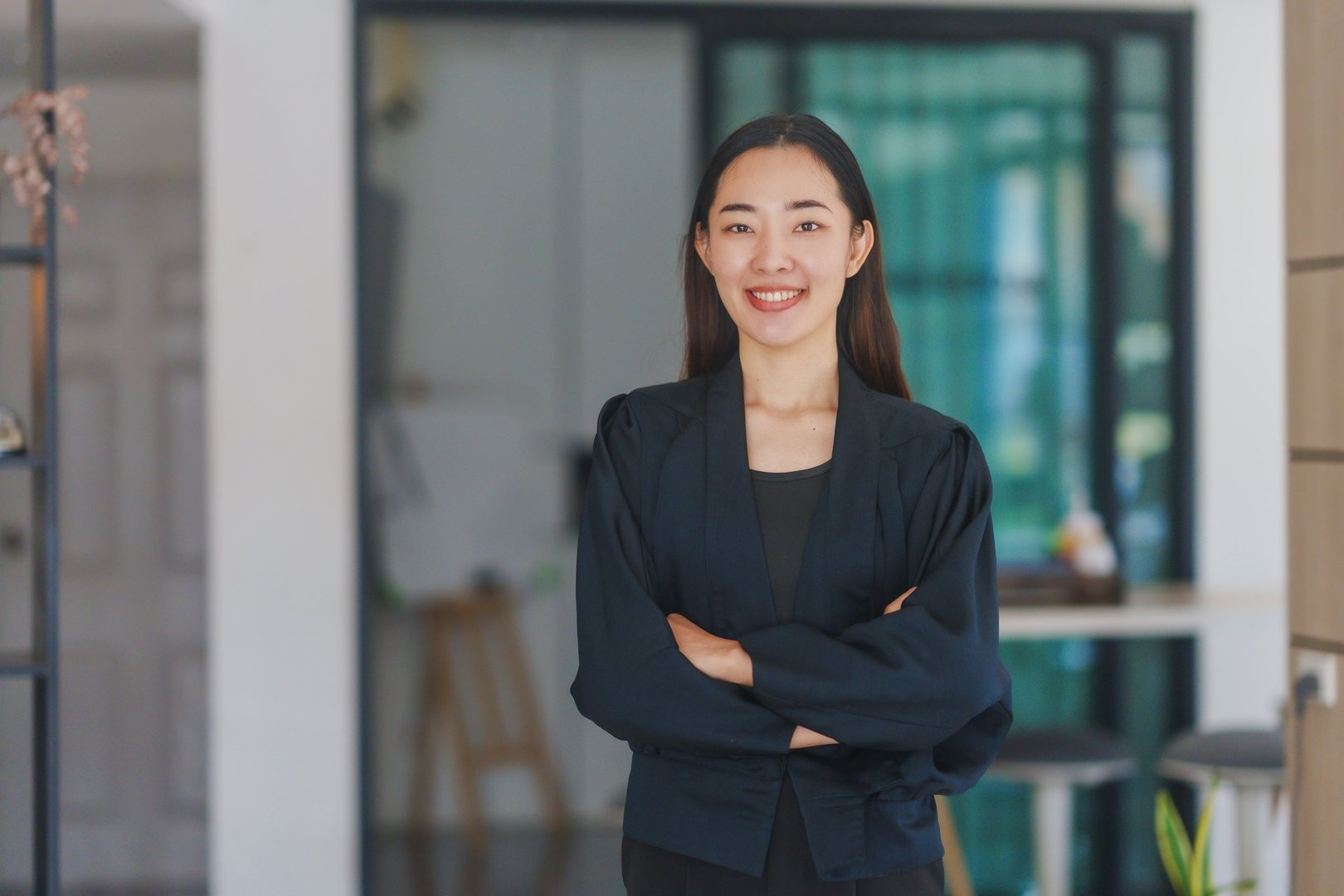 Portrait of a smiling young Asian businesswoman standing with arms crossed in a modern office, exuding confidence and professionalism
