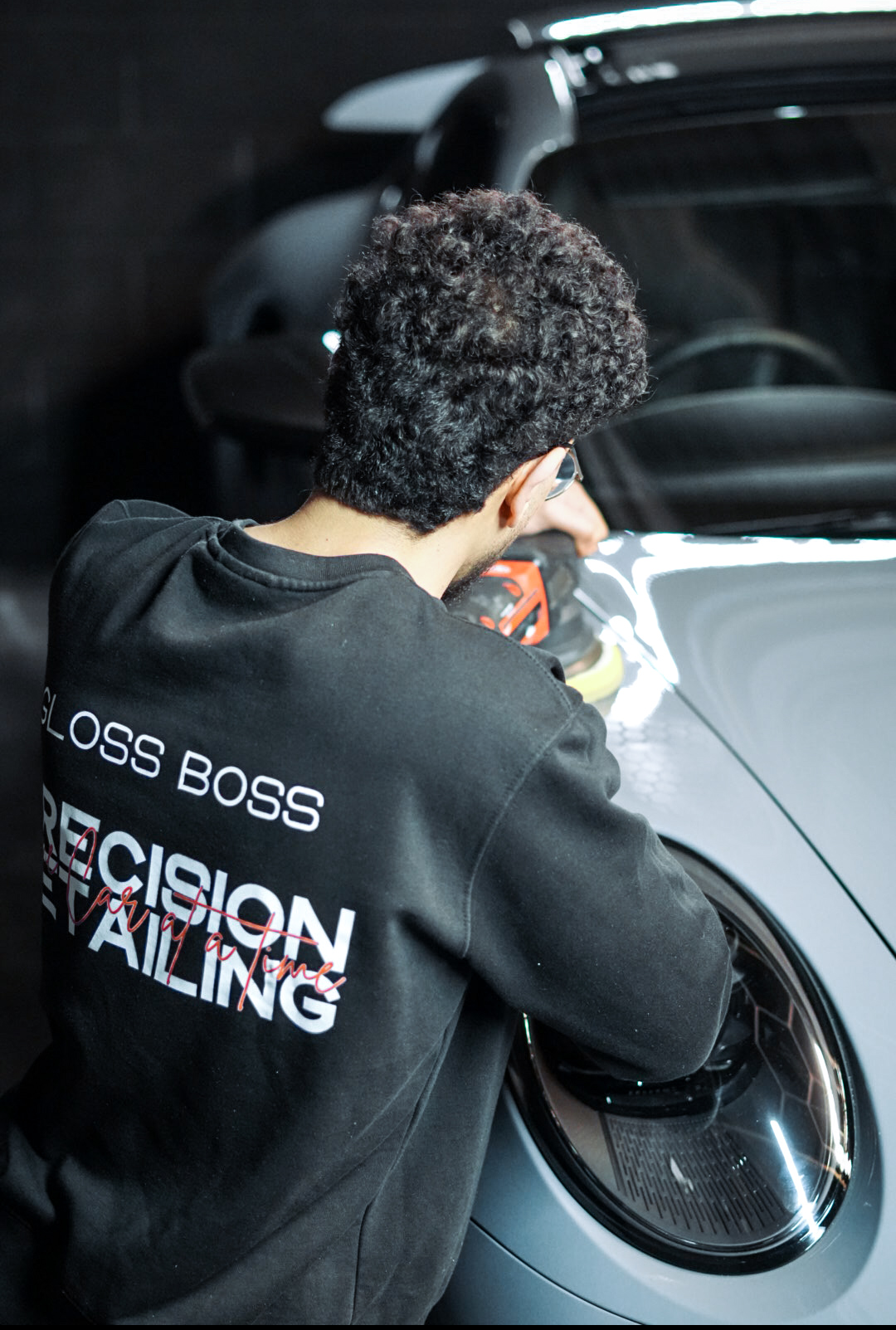 Person polishing the headlight of a silver car, wearing a black Gloss Boss sweatshirt.