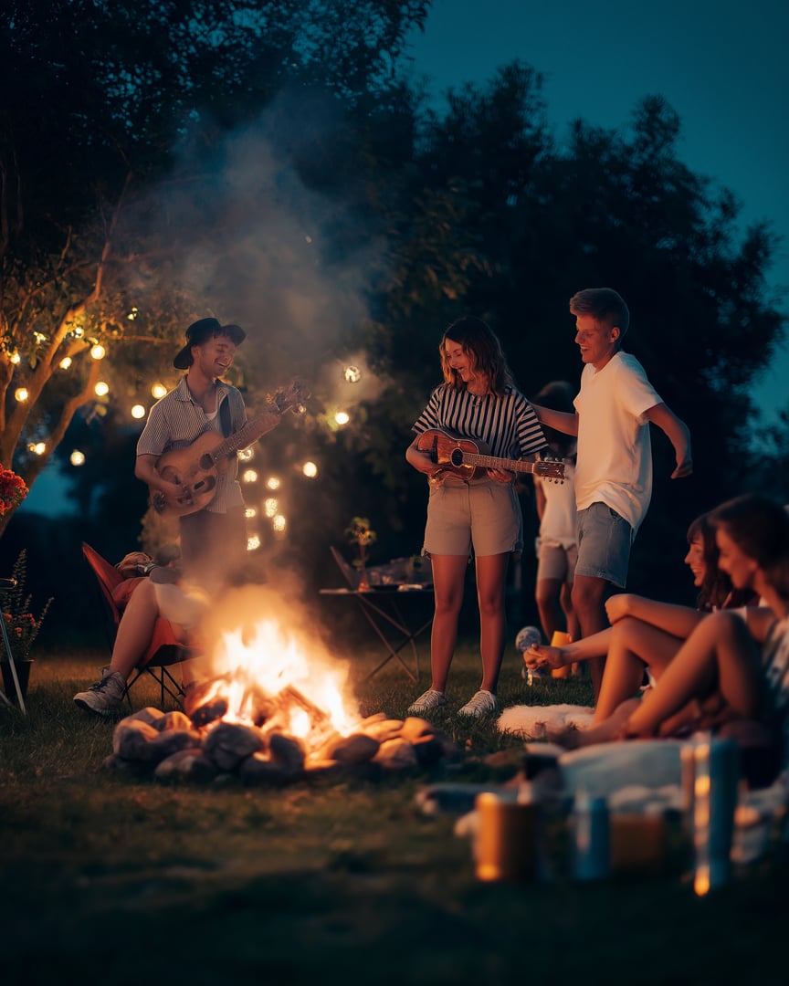People gathered around a campfire, playing guitars and talking, with string lights hanging from trees in the background.