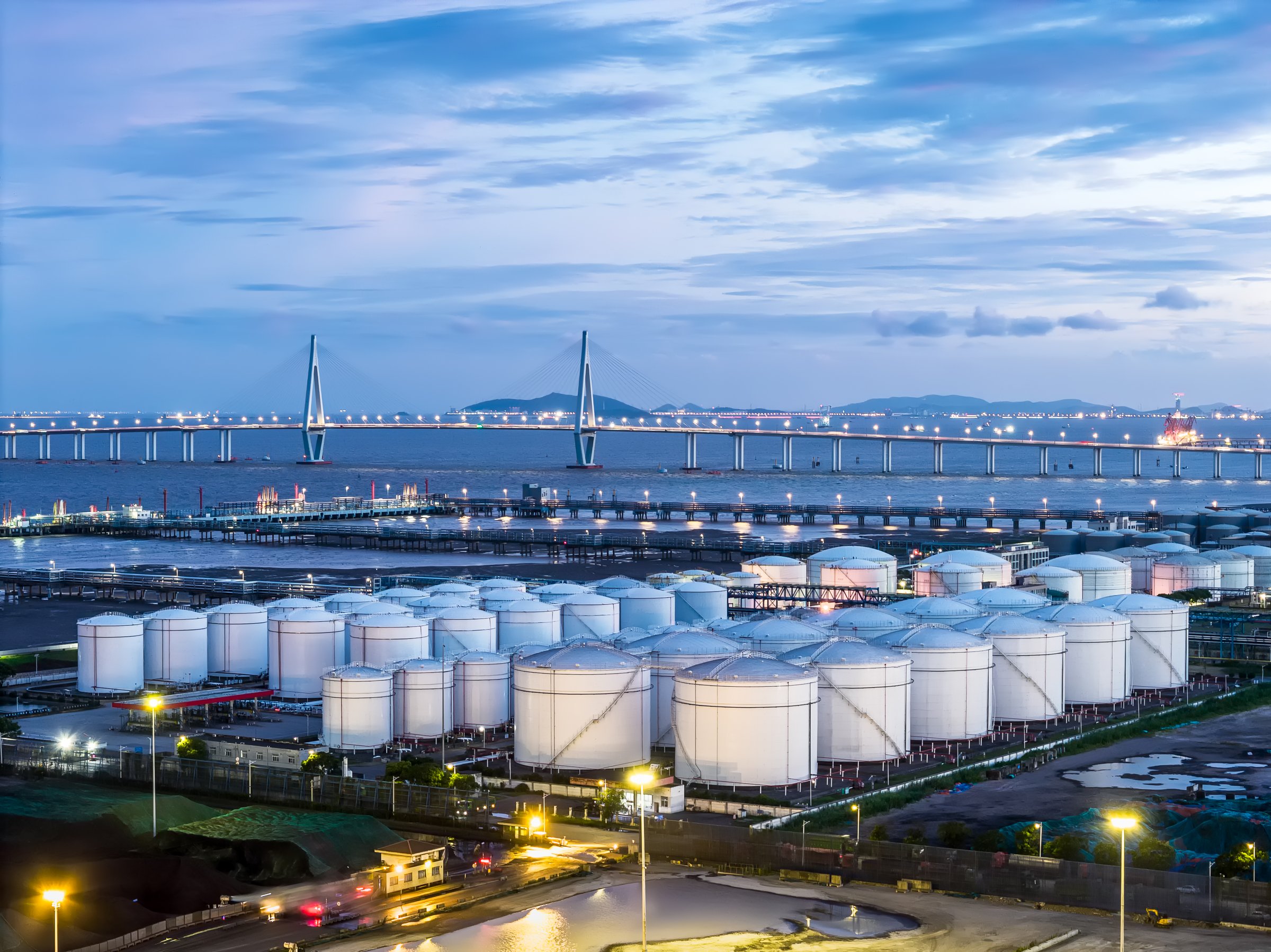 Gleaming white storage tanks of a coastal fuel depot sit near a majestic cable-stayed bridge.