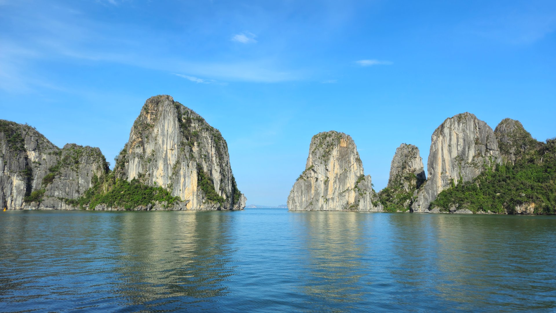 Towering limestone karsts rise from the calm emerald waters of Ha Long Bay, Vietnam, under a clear blue sky.