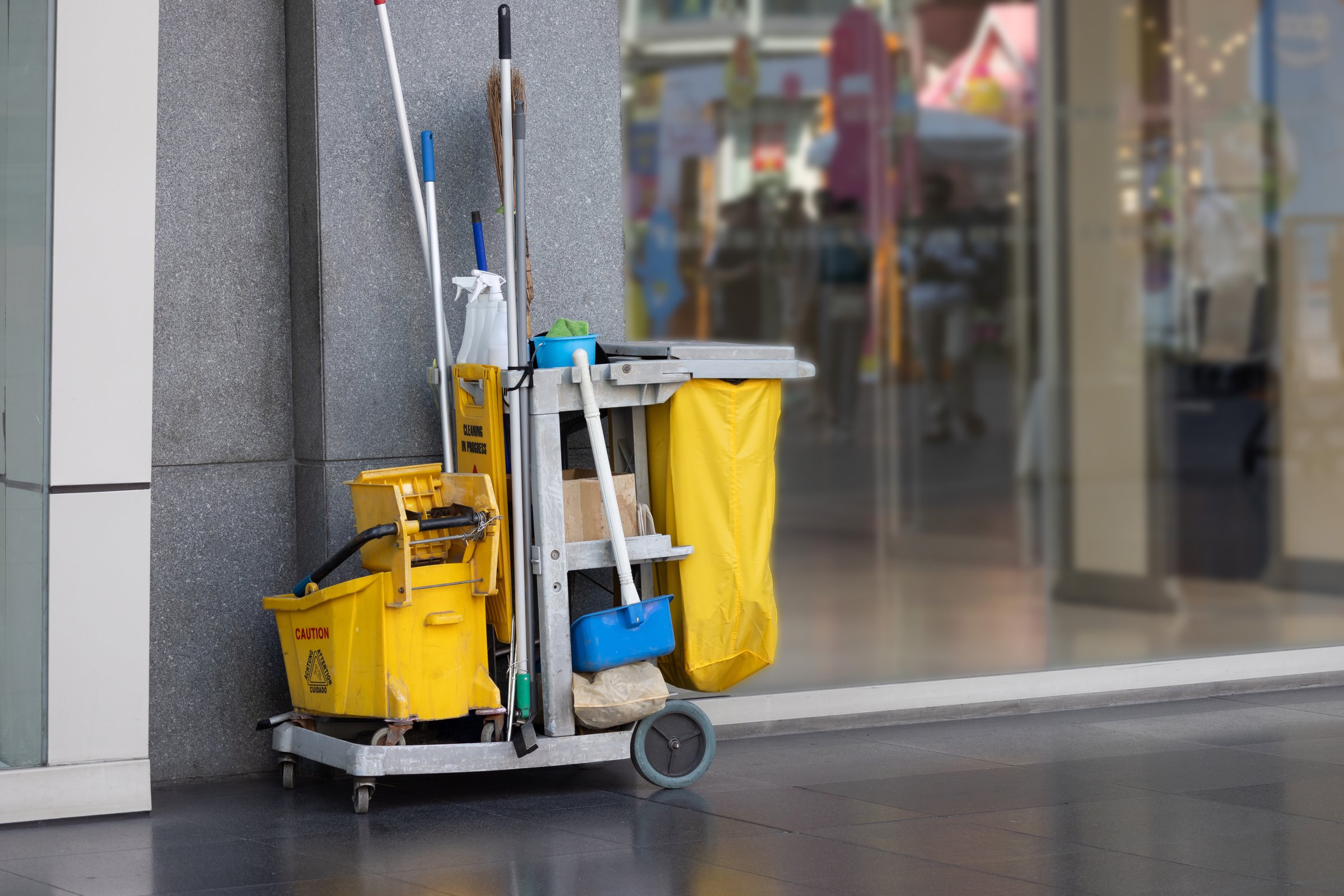 A yellow cleaning cart with a yellow bucket and a blue mop. The cart is parked outside a building