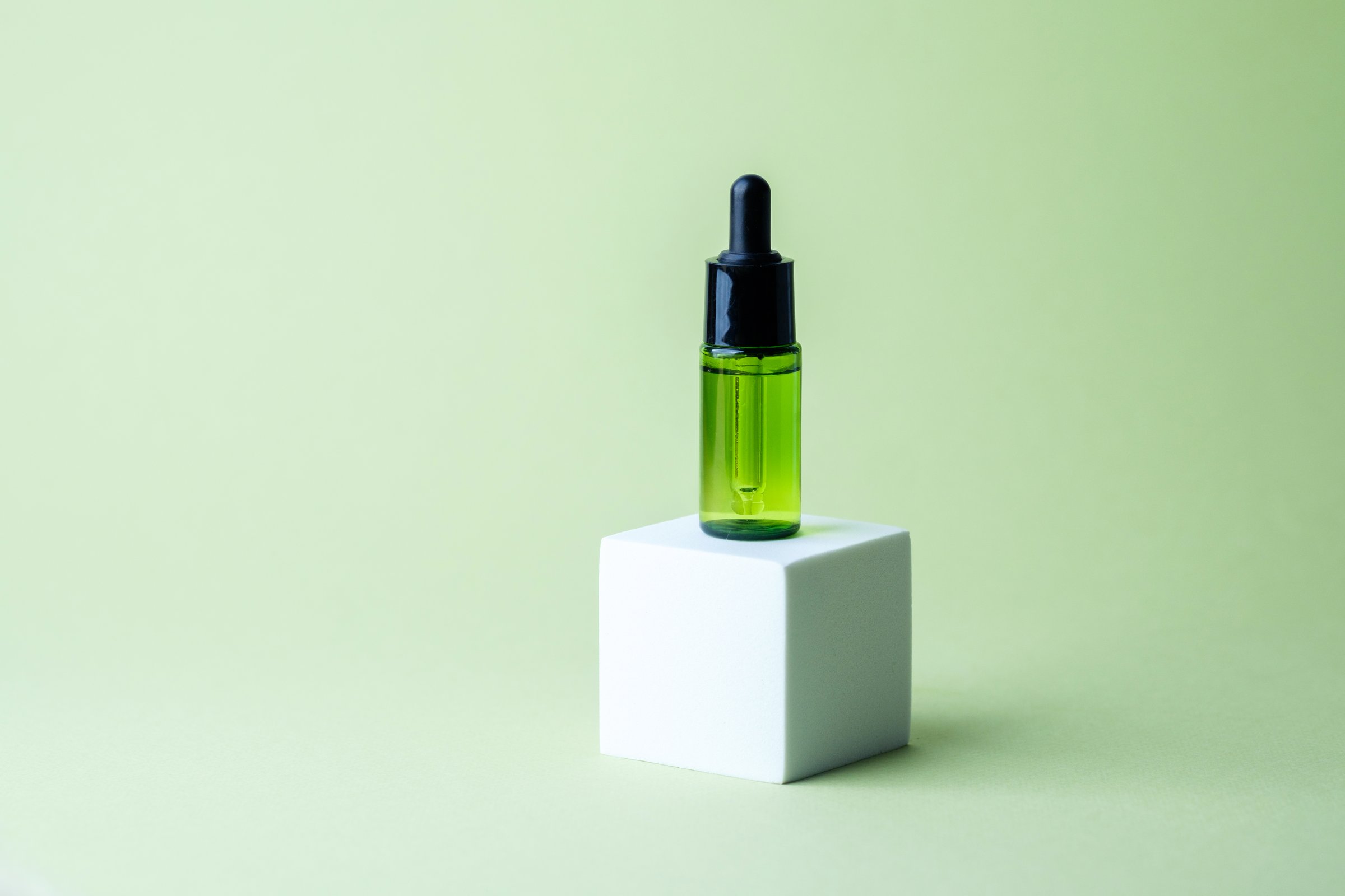 Green cosmetic dropper glass bottle with black cap standing on white cube pedestal on pastel background